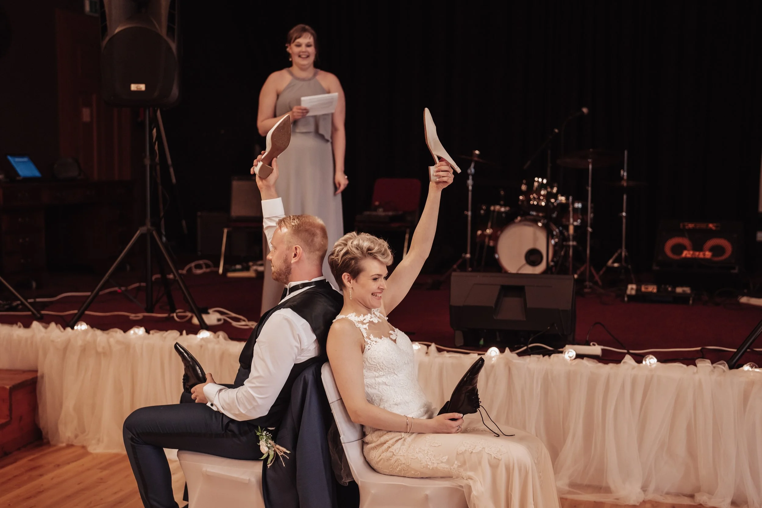 Bride and groom playing shoe game at wedding reception, with female host on stage, and musical equipment in background.