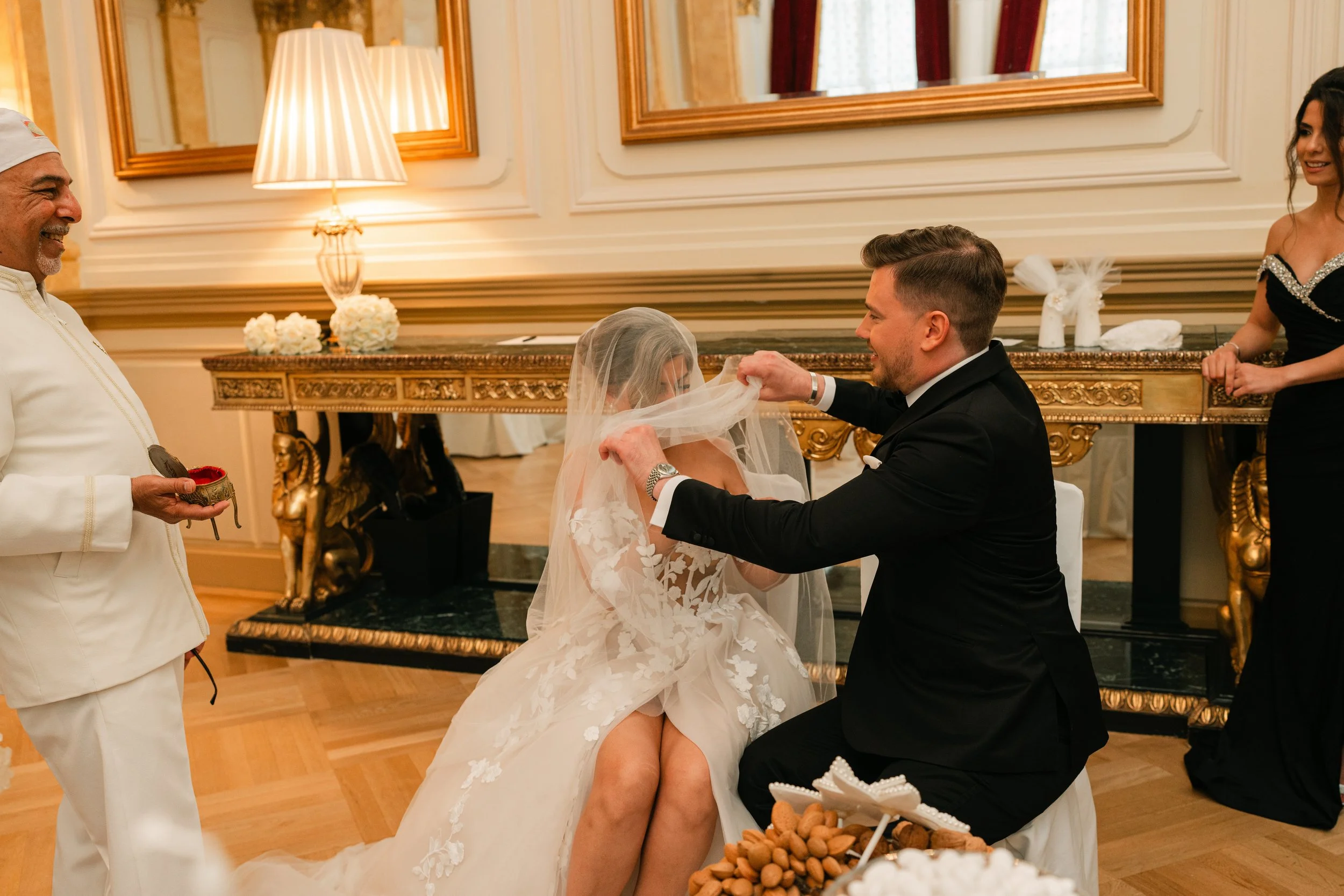 Wedding ceremony with bride and groom, groom lifting bride's veil, man in white holding items, elegant room.