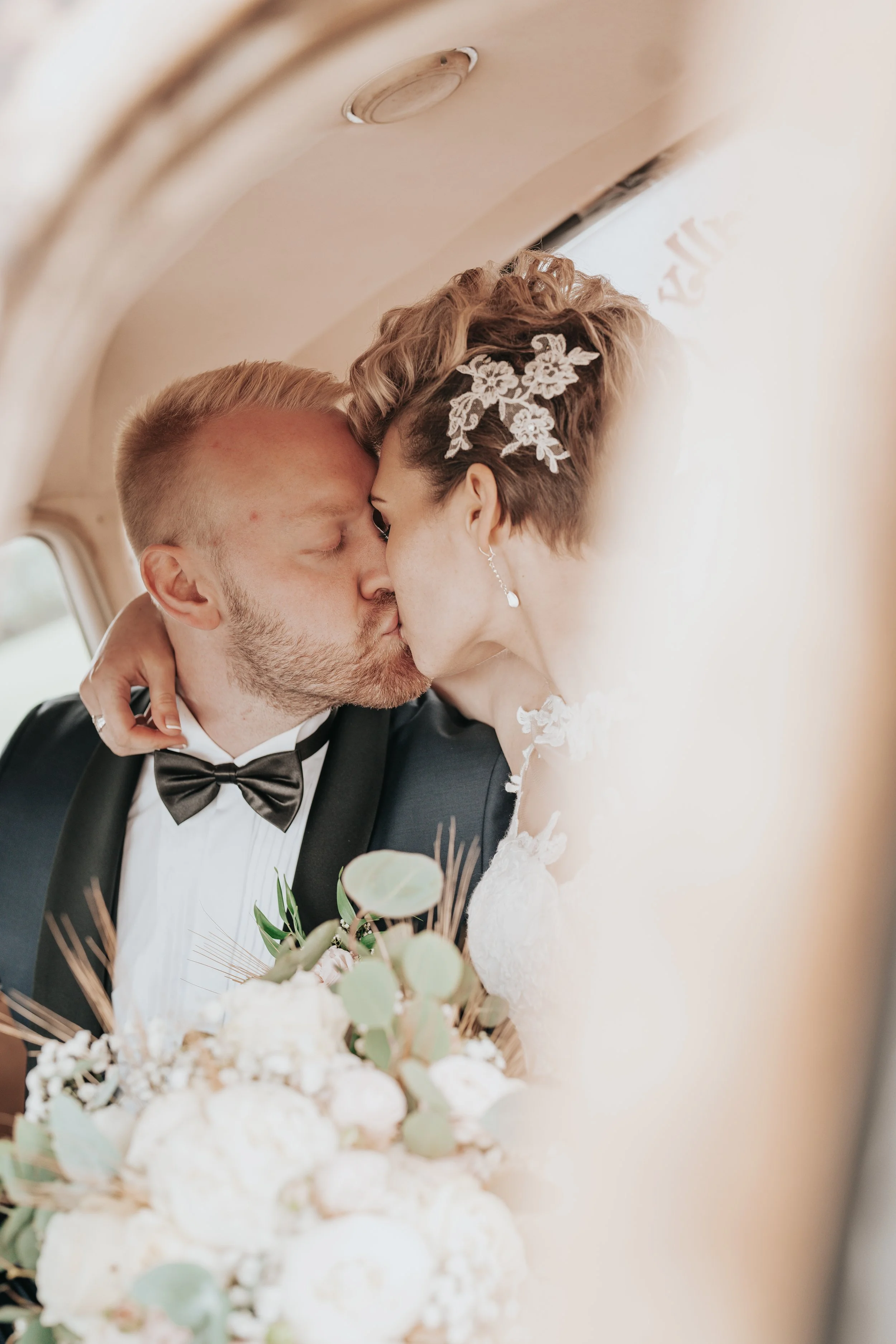 Bride and groom kissing inside a car, bride holding bouquet, groom wearing a bow tie.