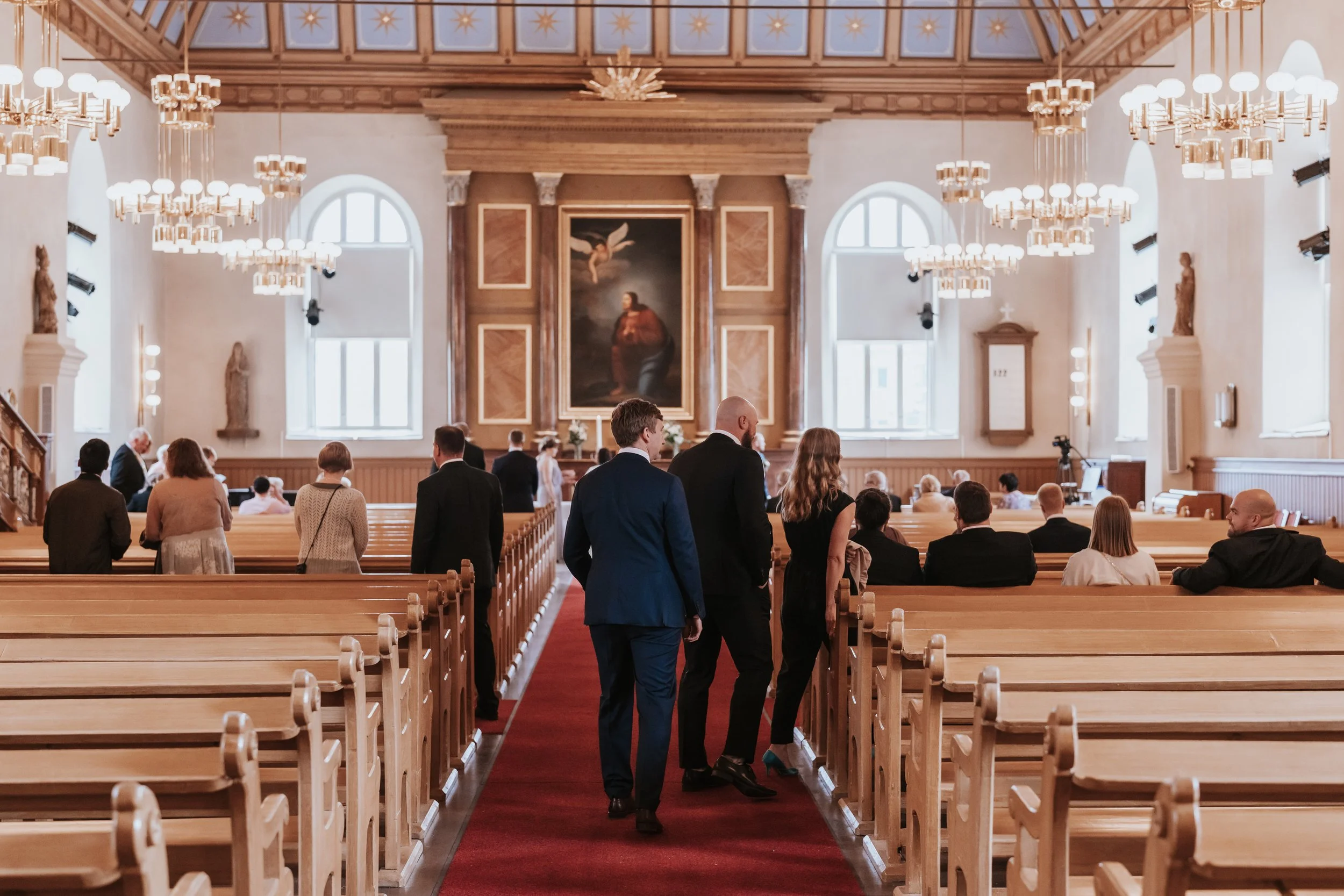 Interior of a church with people sitting and standing, chandeliers hanging from ceiling, and religious artwork on the wall.