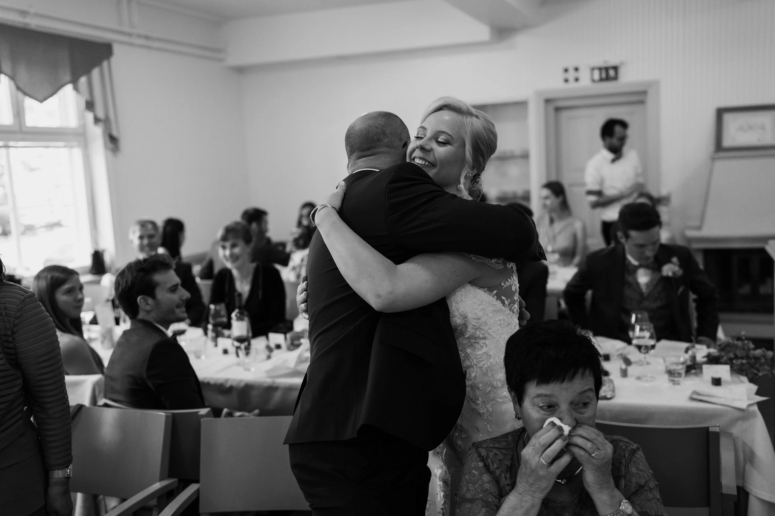 Wedding reception, bride hugging a guest, woman wiping tears in foreground, guests seated at tables, black and white photo.
