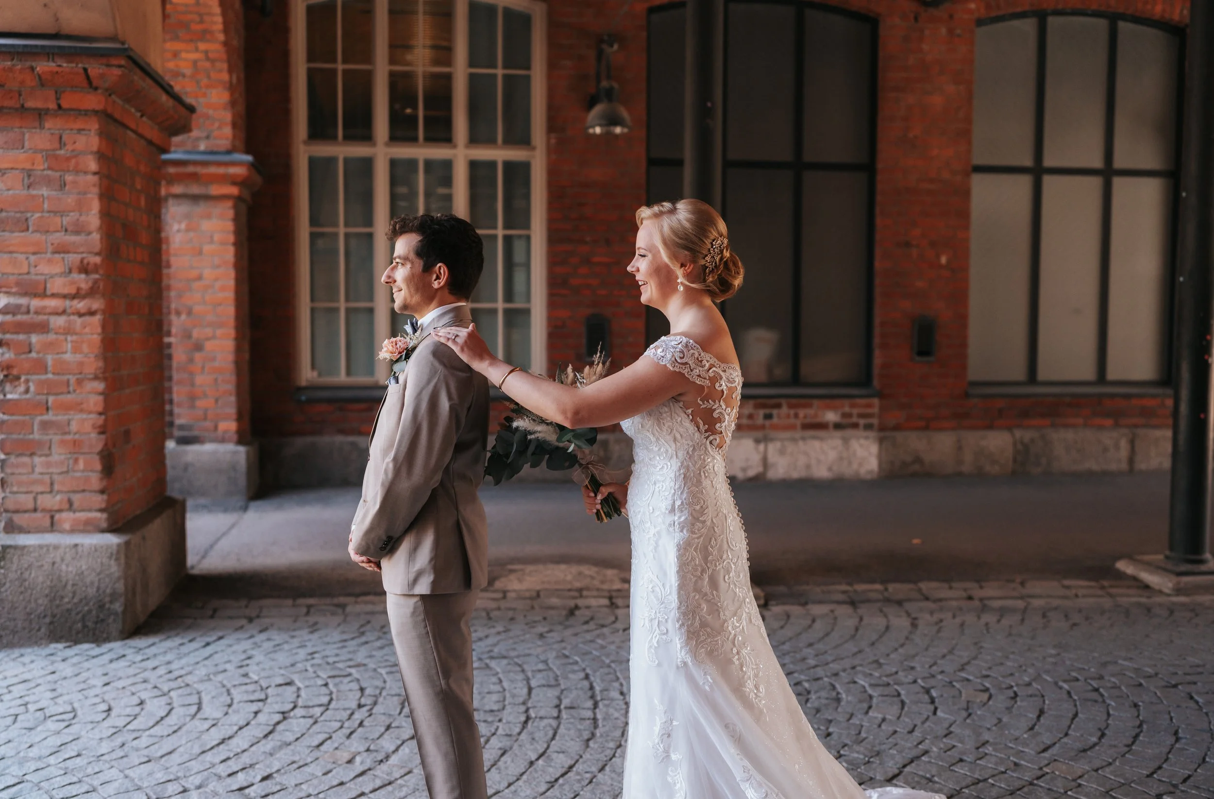 Bride in a lace wedding dress surprises groom in beige suit, standing in front of a brick building.