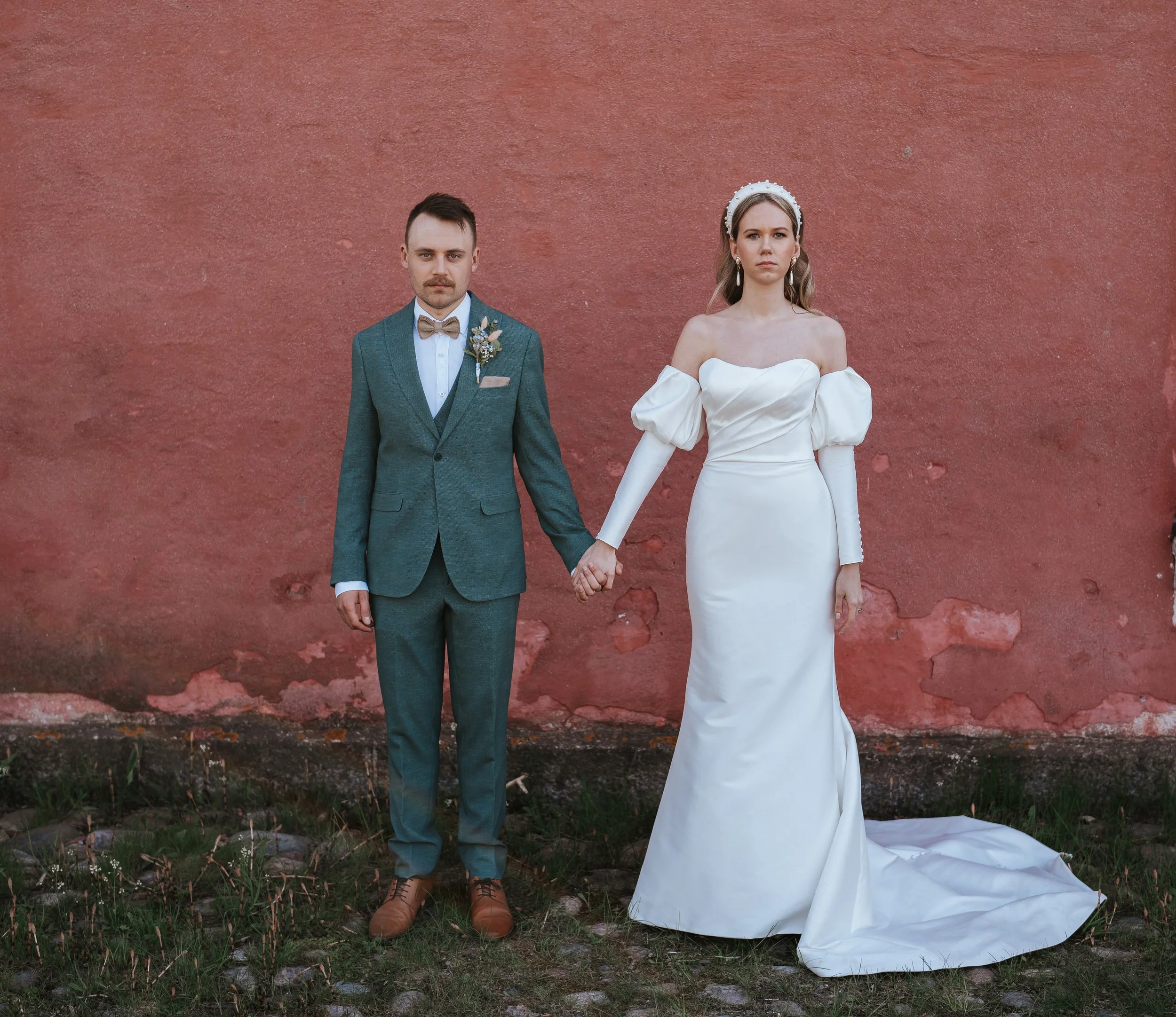 Bride in white gown and groom in teal suit holding hands against a red wall backdrop.