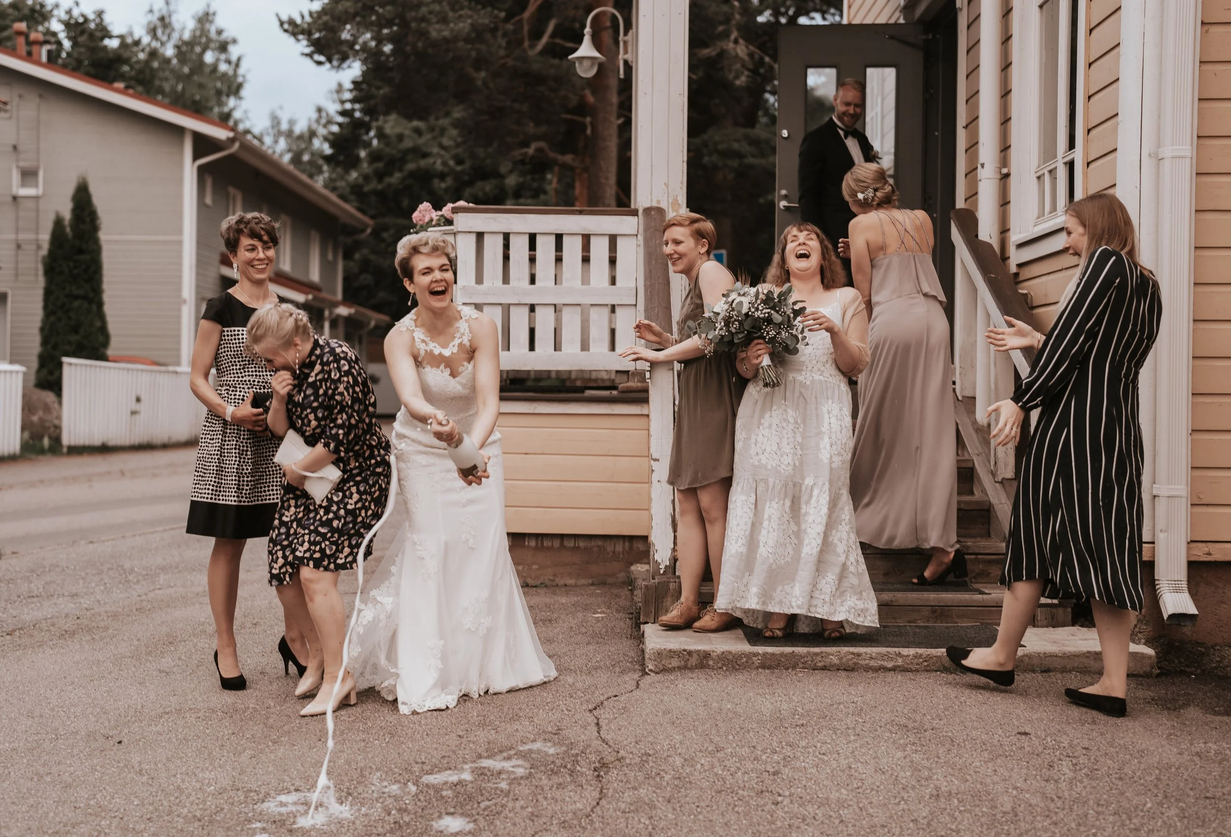 Group celebrating outside, woman in wedding dress popping champagne, people laughing, outdoor scene, candid moment.