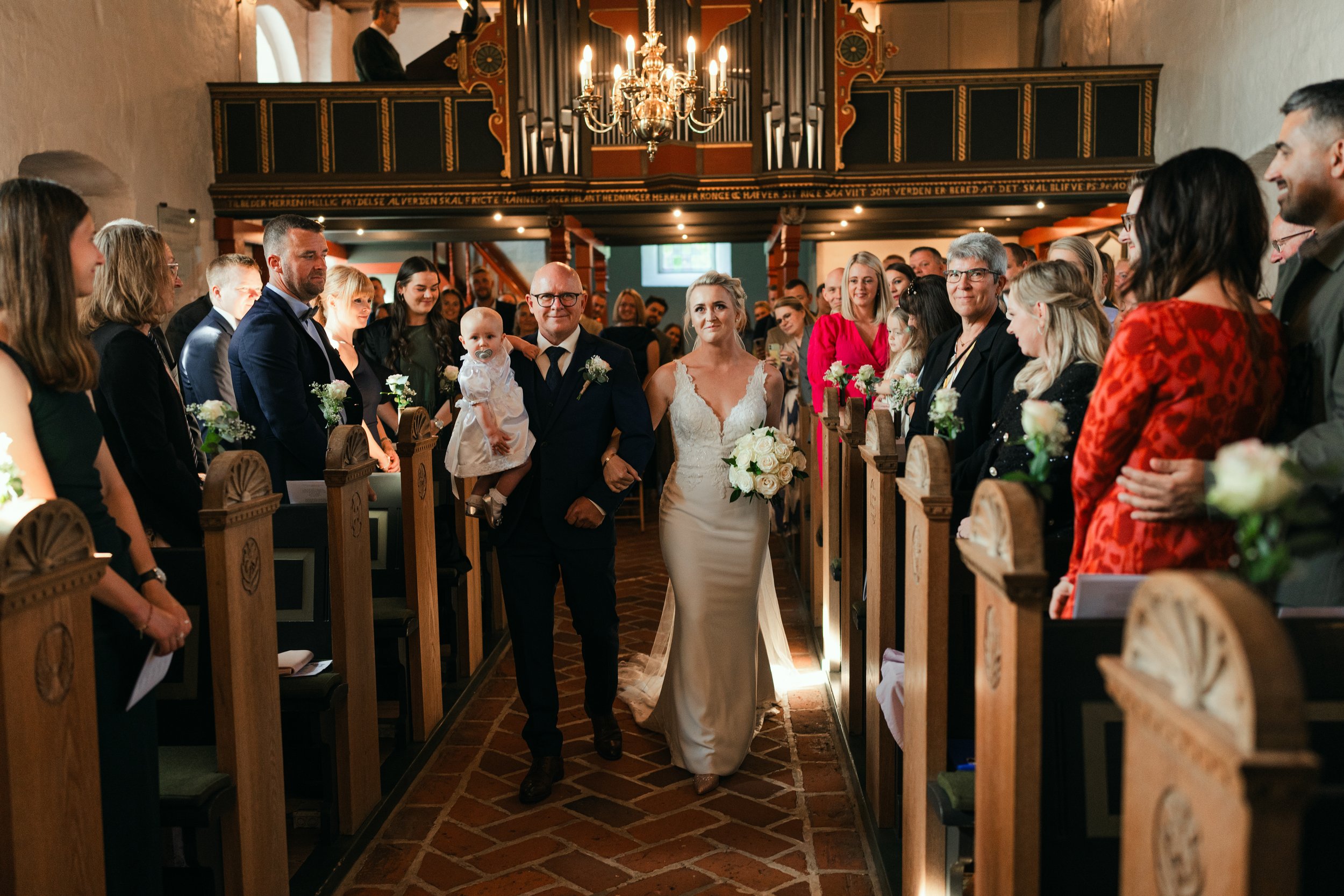 Bride walking down the aisle with an older man holding a baby in a church setting, surrounded by guests, flowers on pews, and a chandelier overhead.