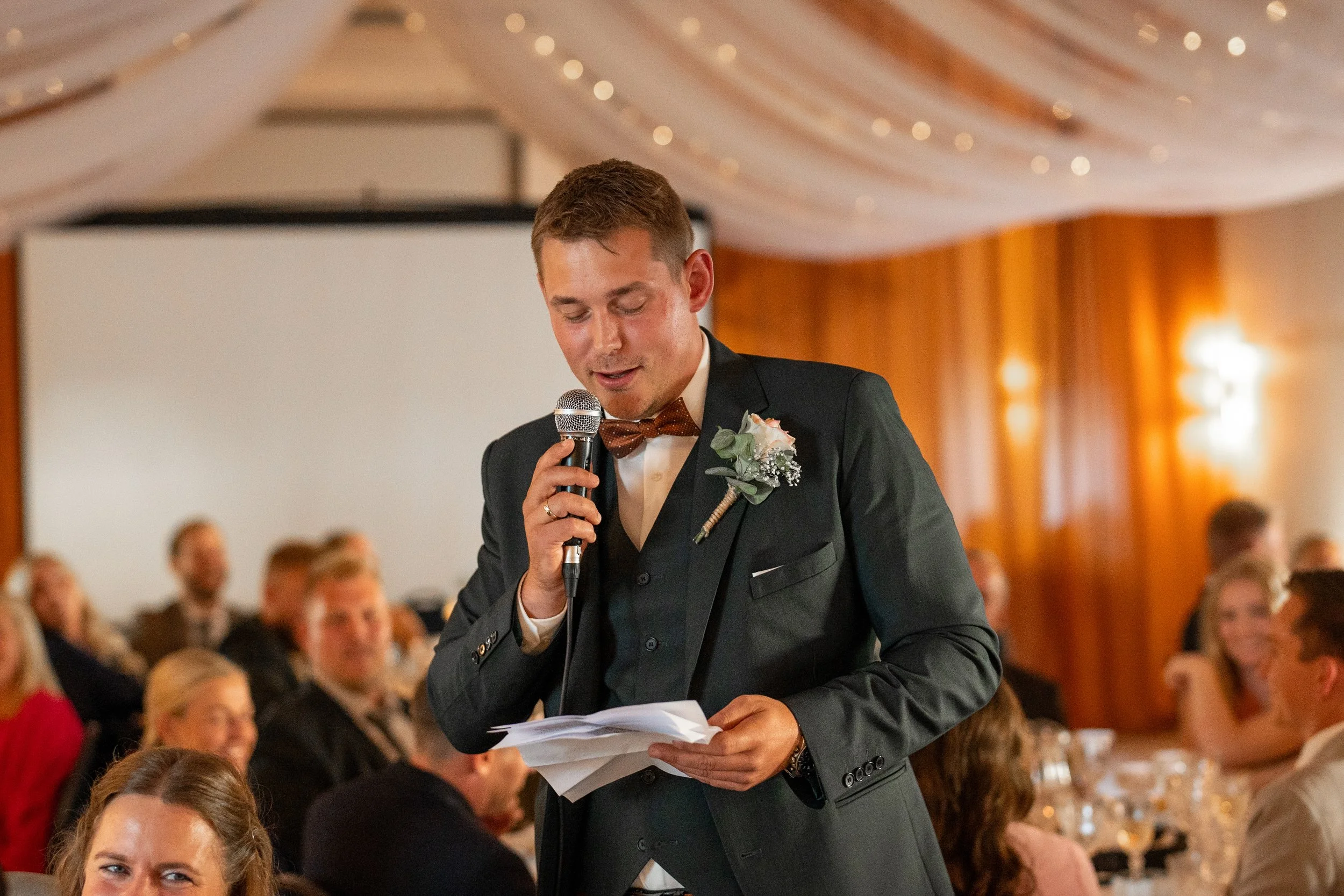 Man in suit giving a speech at a wedding reception with microphone and notes, surrounded by guests.