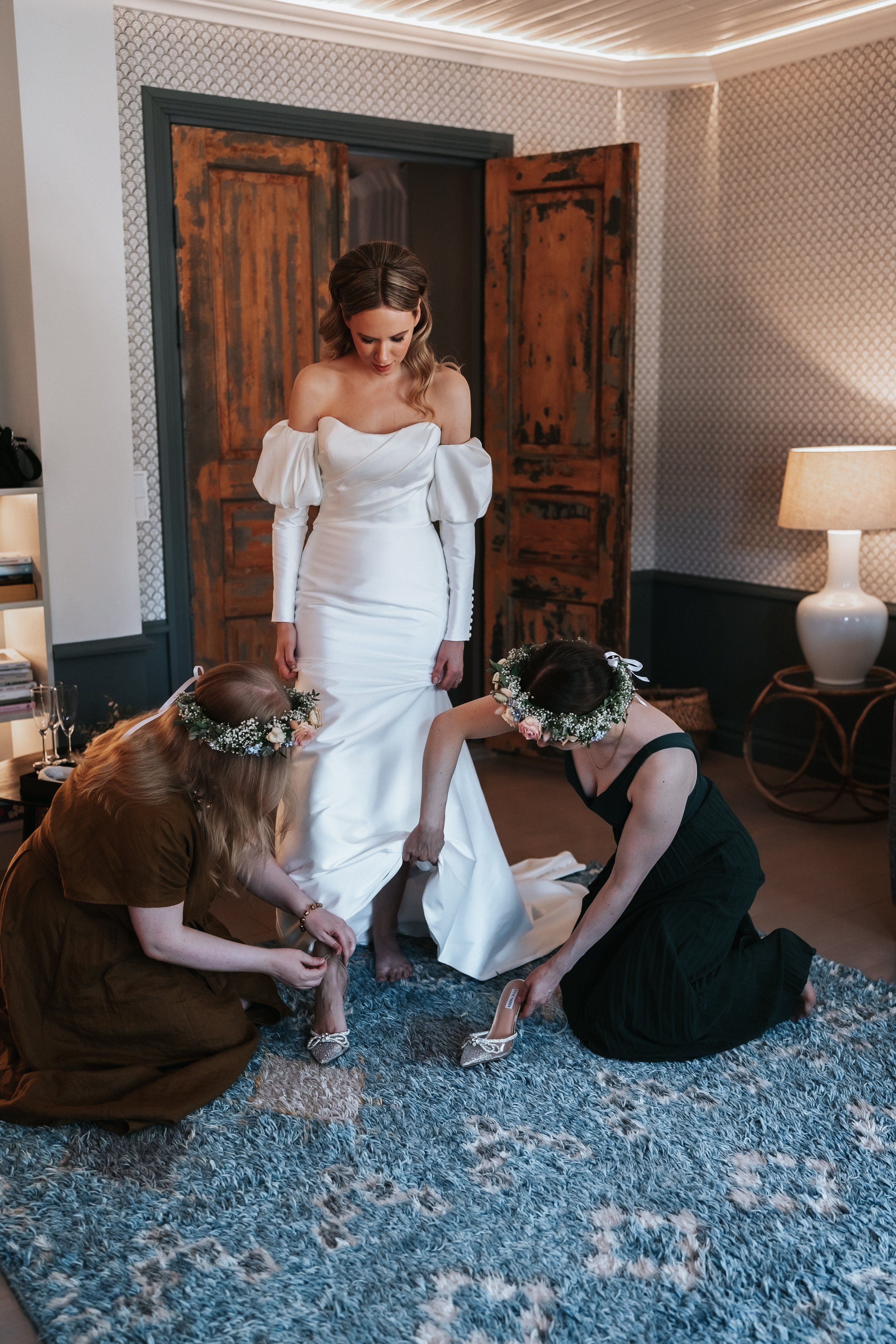 Bride in white wedding dress being assisted by two women on a blue carpet in a room with wooden doors.