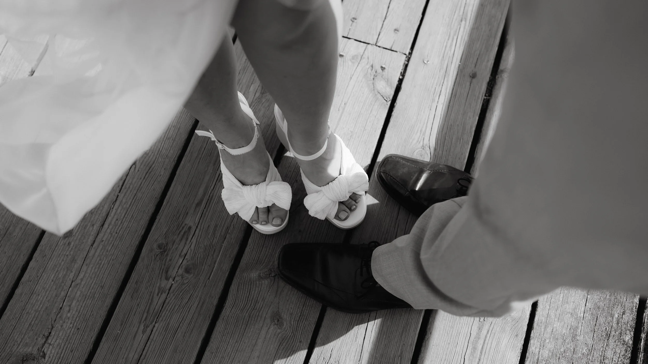 Close-up of a bride's white shoes next to black shoes on a wooden floor