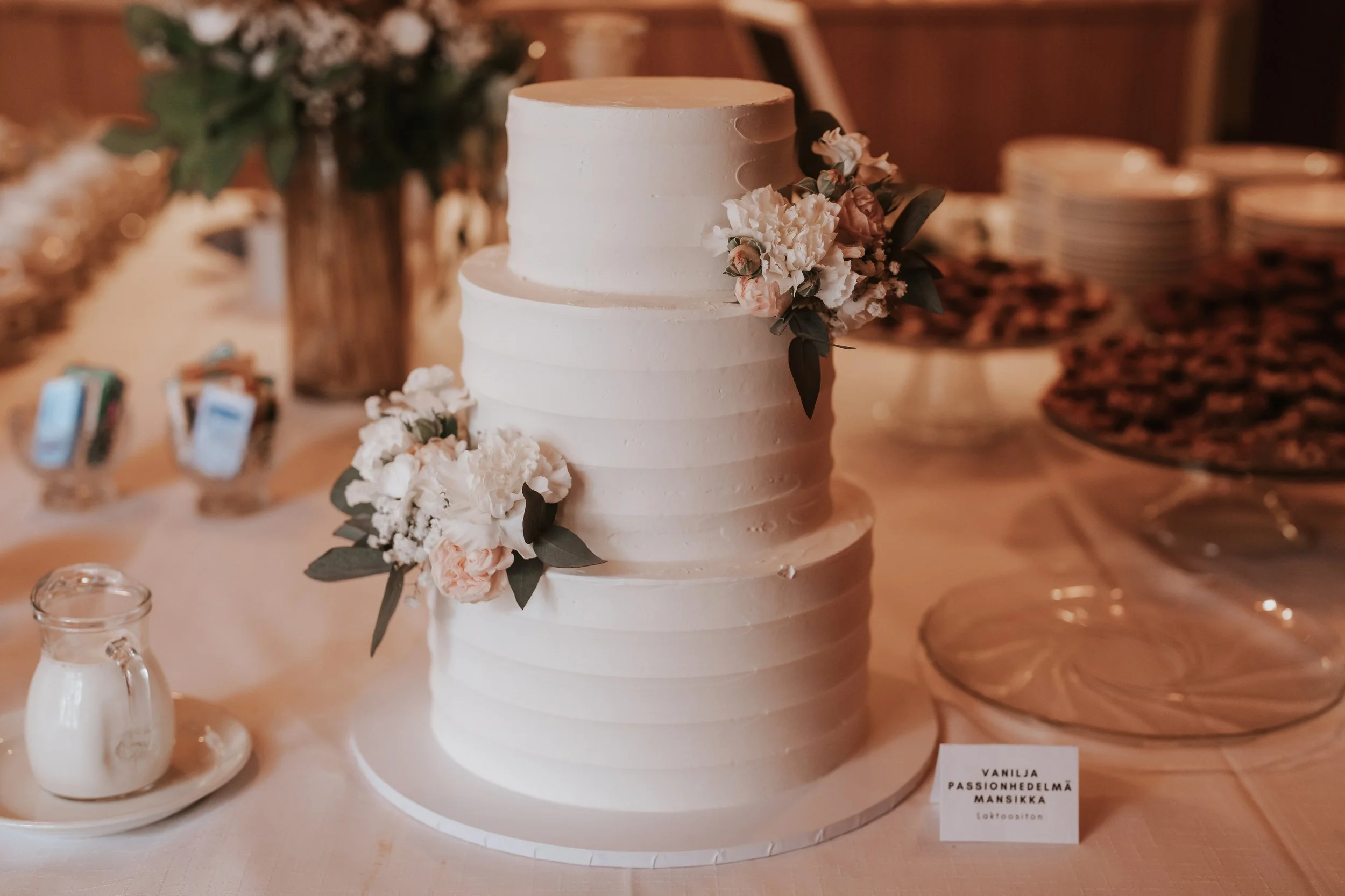 Three-tier white wedding cake with floral decorations, placed on a table with desserts and a jar of cream.