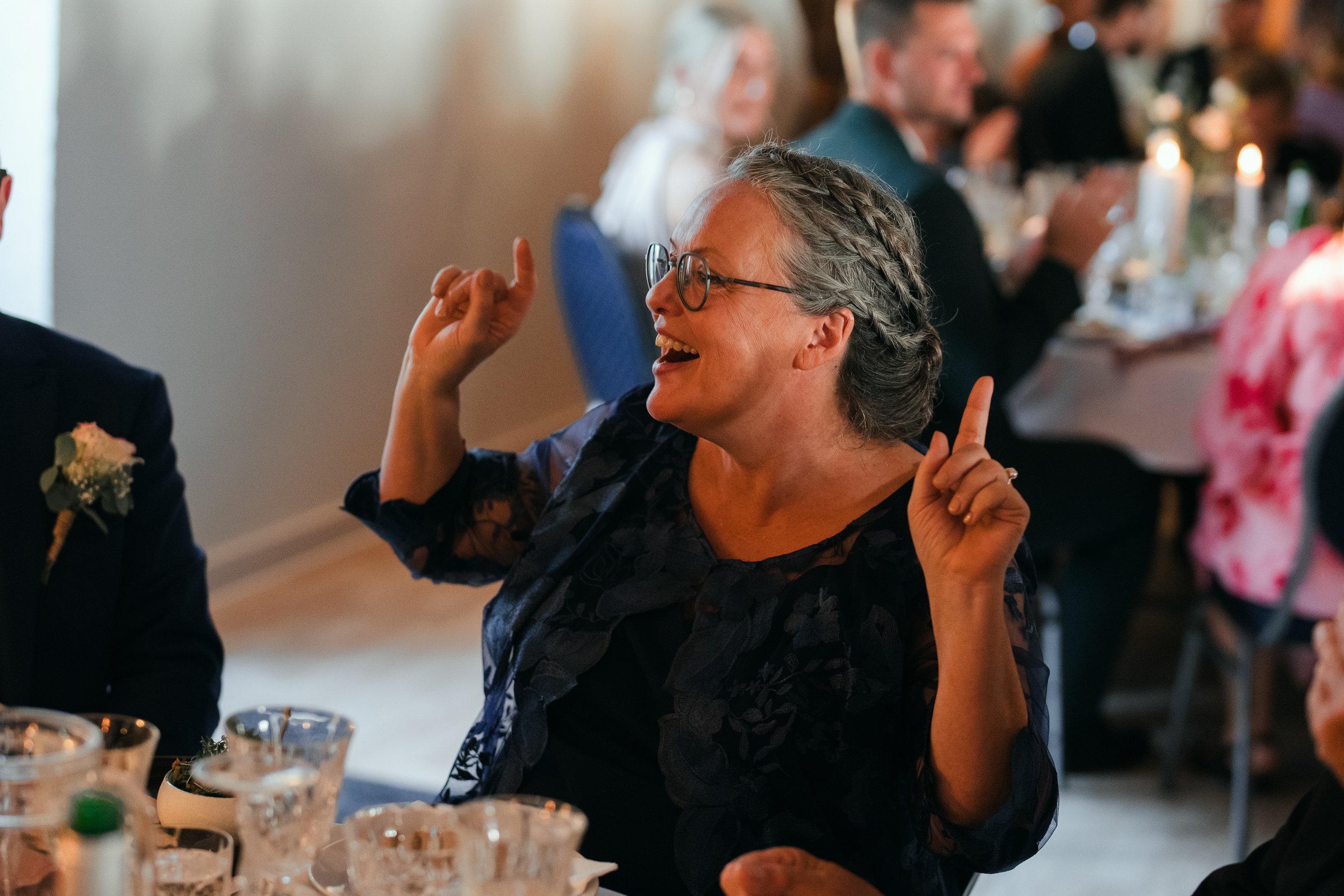 Woman smiling and gesturing with her hands at a social event, seated at a table with candles and glassware in the background.