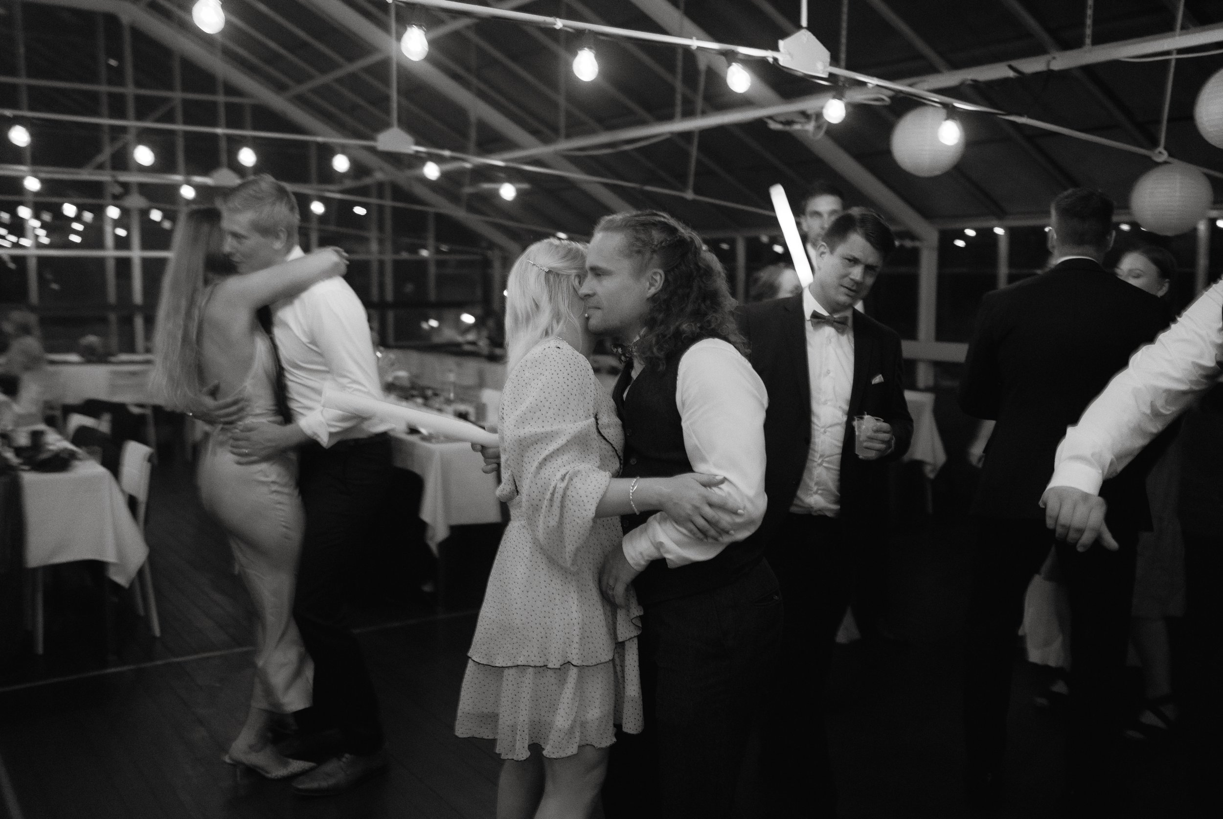 Black and white photo of people dancing at an indoor event with hanging lights and tables.