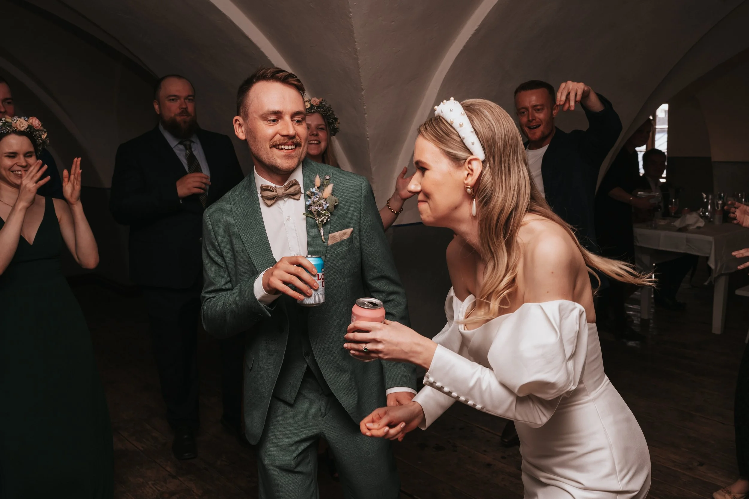 Wedding celebration with bride in white dress and groom in green suit holding drinks, surrounded by guests clapping and dancing in a dimly lit room.