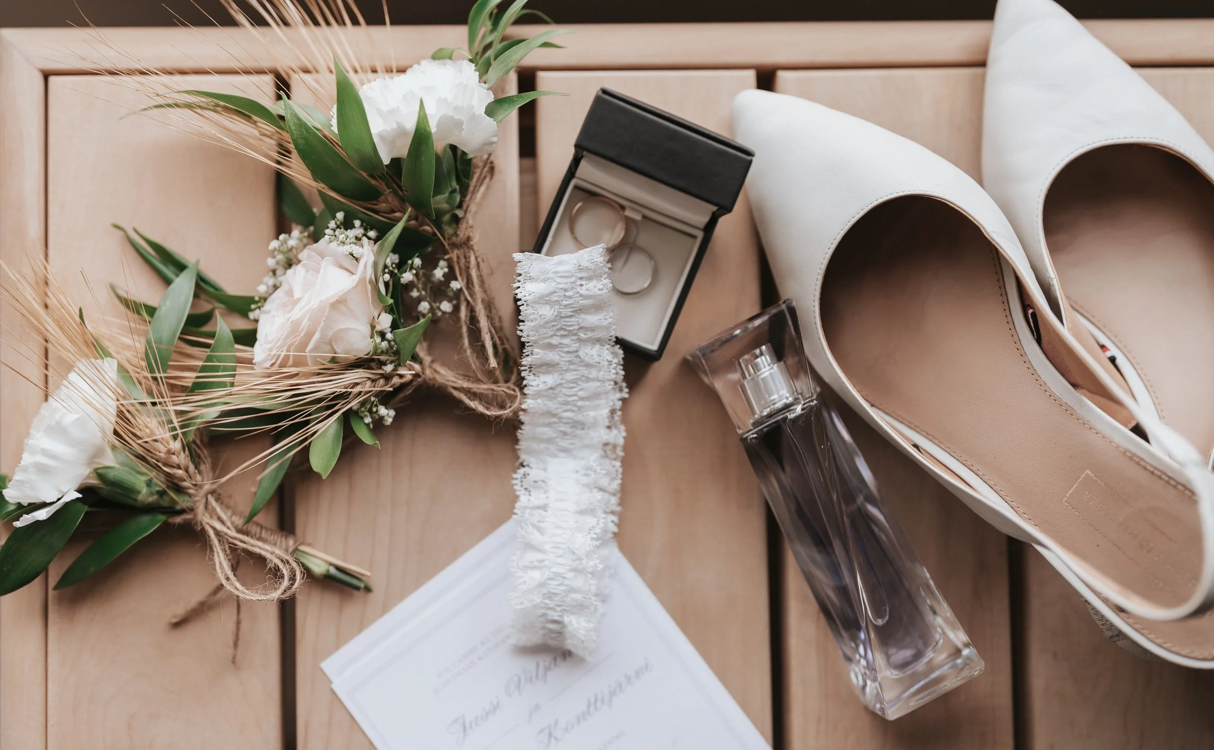 Flat lay of wedding items including white heels, a garter, wedding rings in a box, a perfume bottle, floral boutonniere, and an invitation card on a wooden surface.