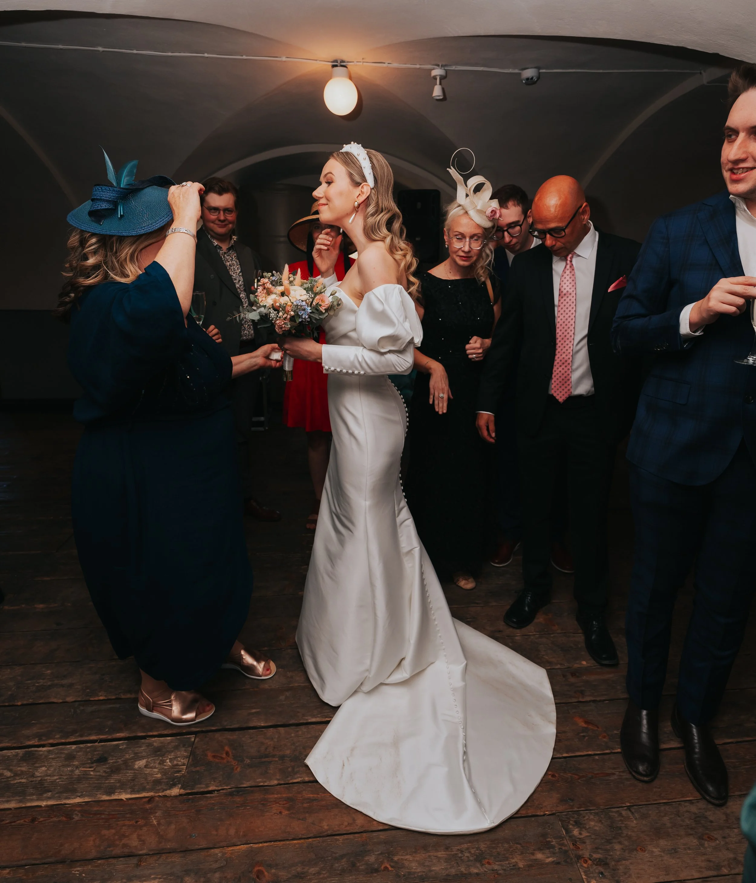 Bride in a white wedding dress surrounded by guests at a reception, holding a bouquet, indoors with wooden flooring and dim lighting.