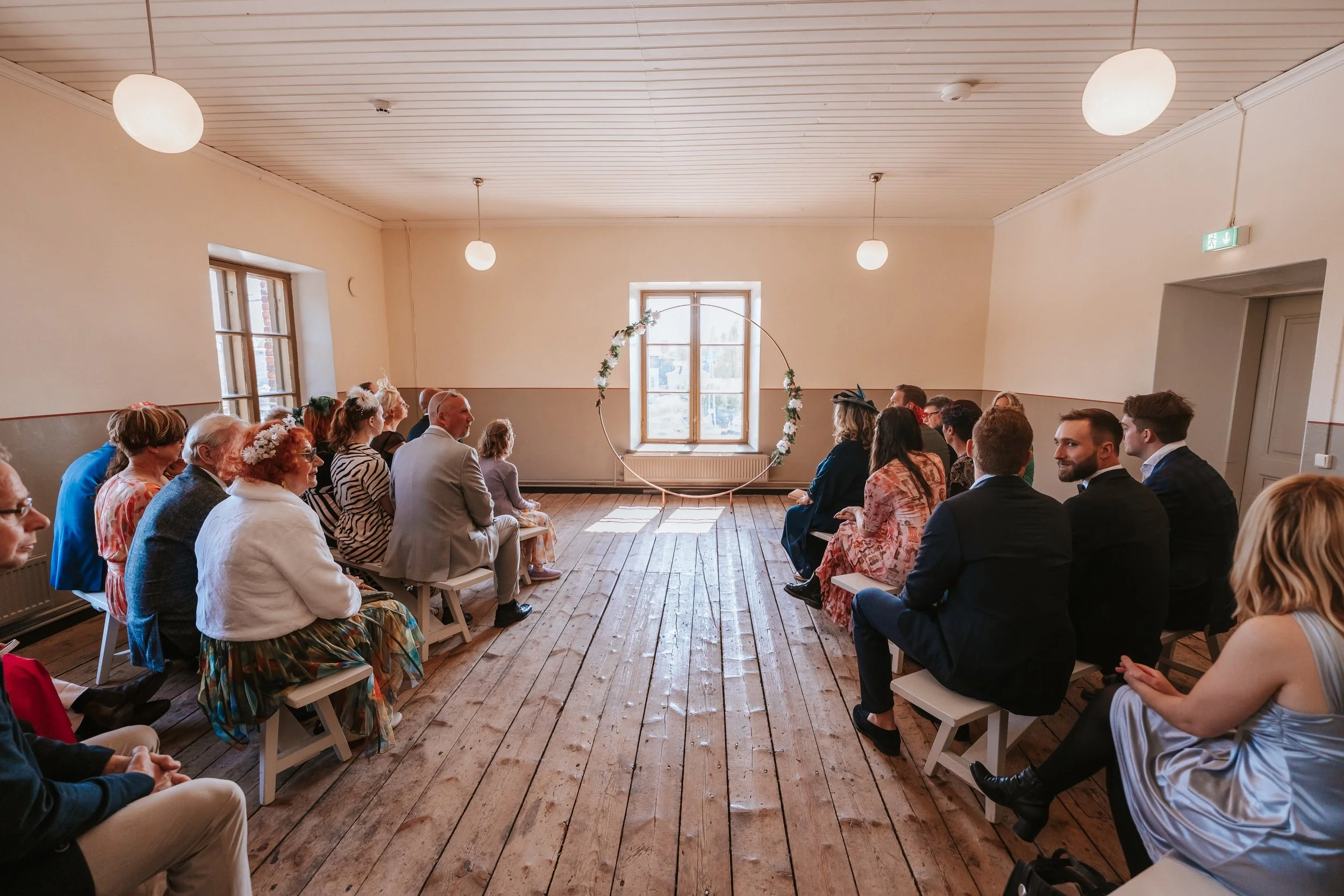 Indoor wedding ceremony setup with guests seated on benches, facing a floral-decorated circular arch in front of a window.