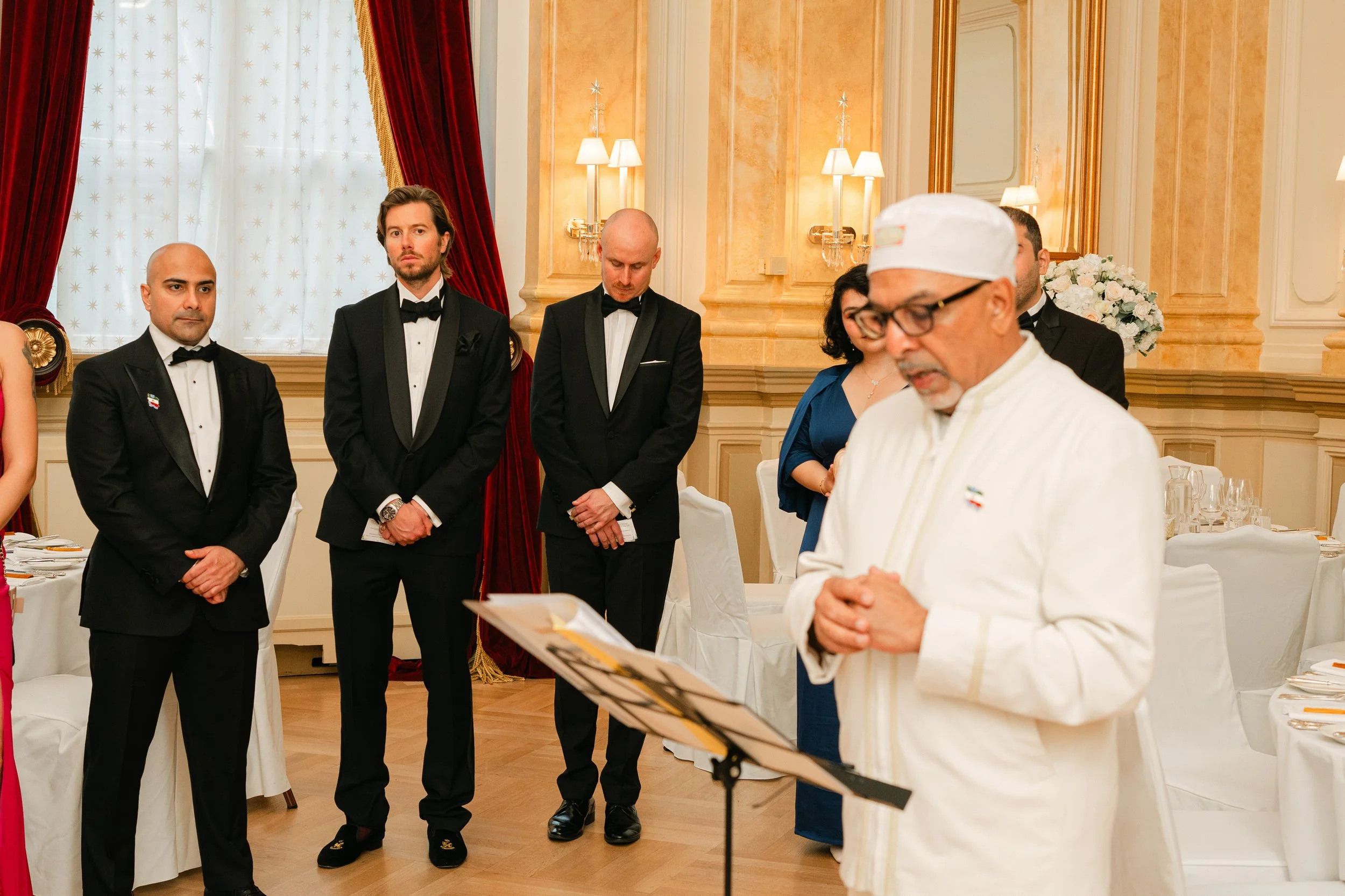 A group of people in formal attire at a ceremonial event with a man in white leading, standing in an elegant room with red curtains and ornate decor.