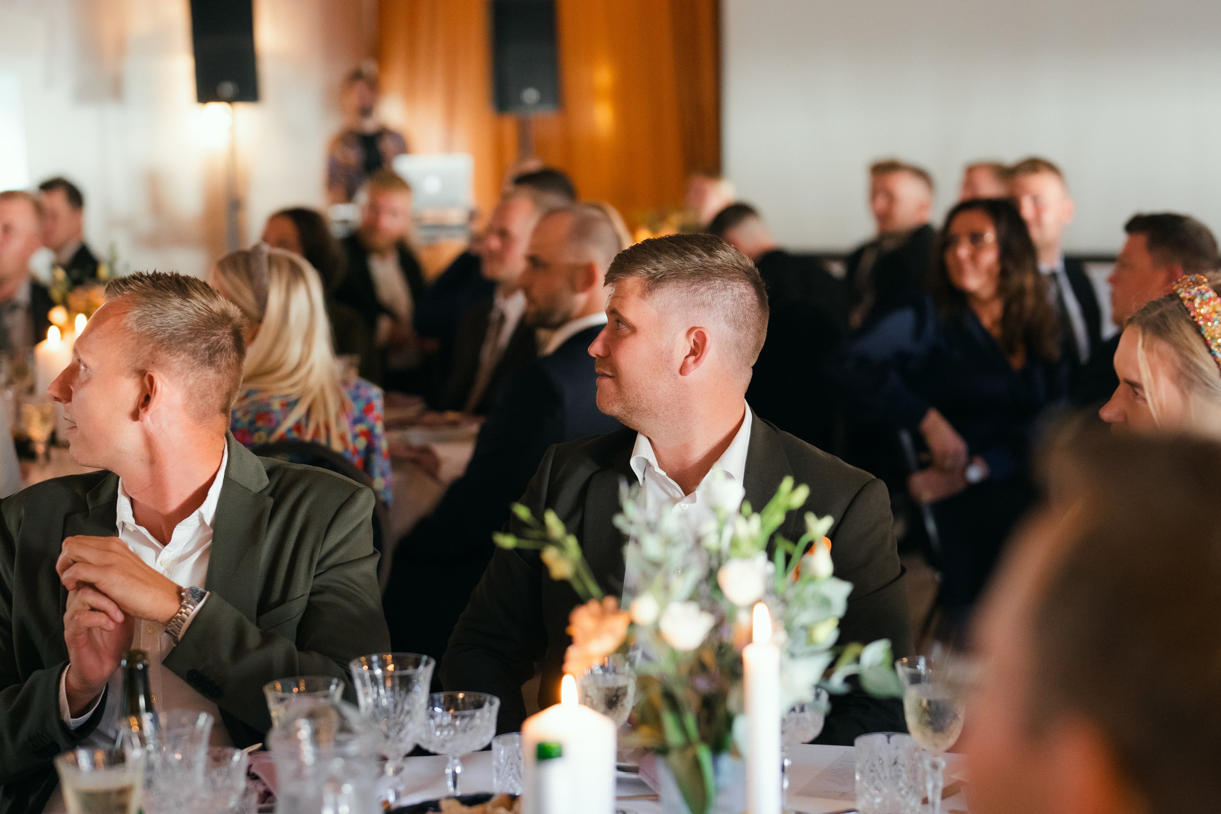 People seated at a formal event with tables set for dining, candles and floral centerpieces, and a speaker in the background.