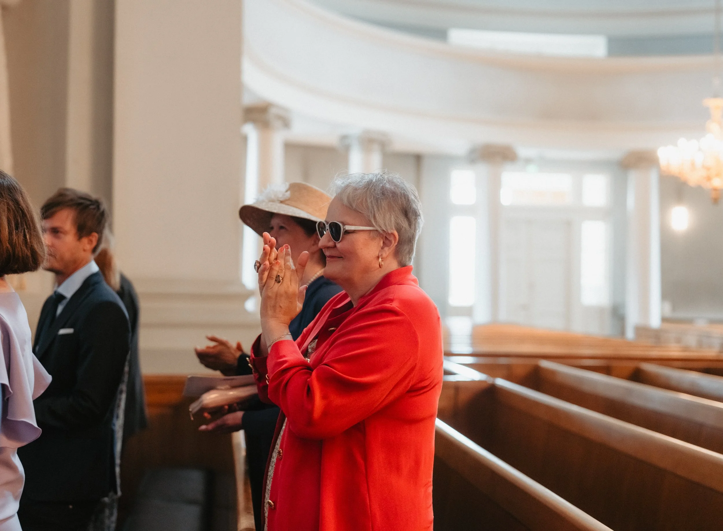 People in church wearing formal attire, including a woman in a red coat clapping.