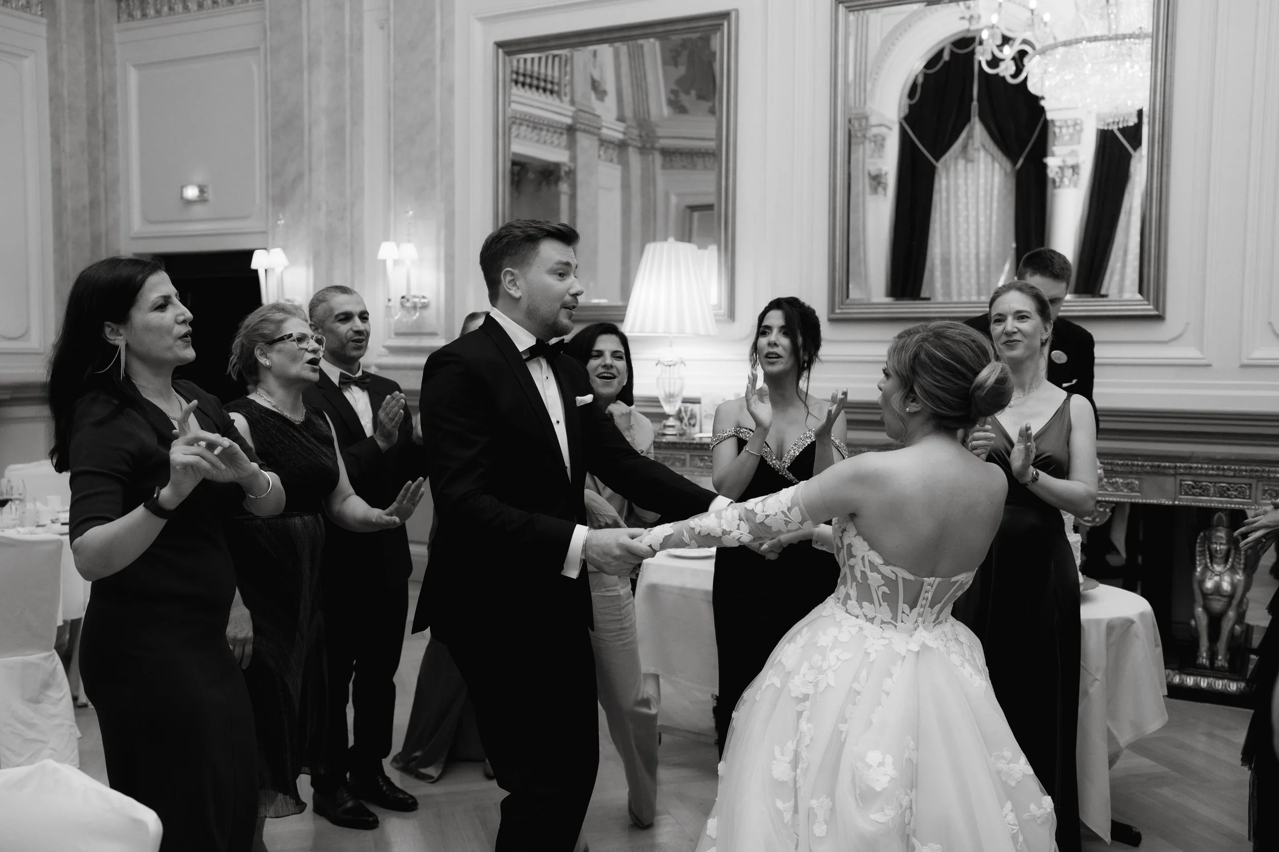 Black and white photo of people dancing at an elegant indoor event. The bride in a wedding dress is dancing with a man in a tuxedo, surrounded by others clapping and smiling.