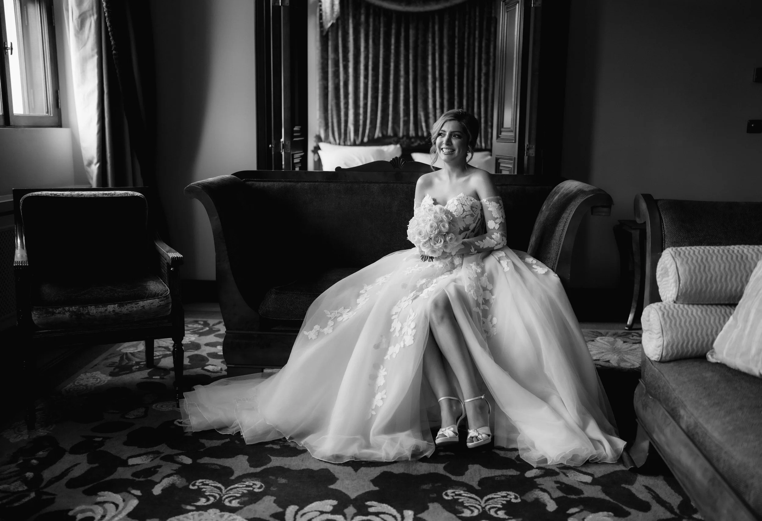 Black and white image of a bride in a wedding dress sitting on a couch, holding a bouquet.