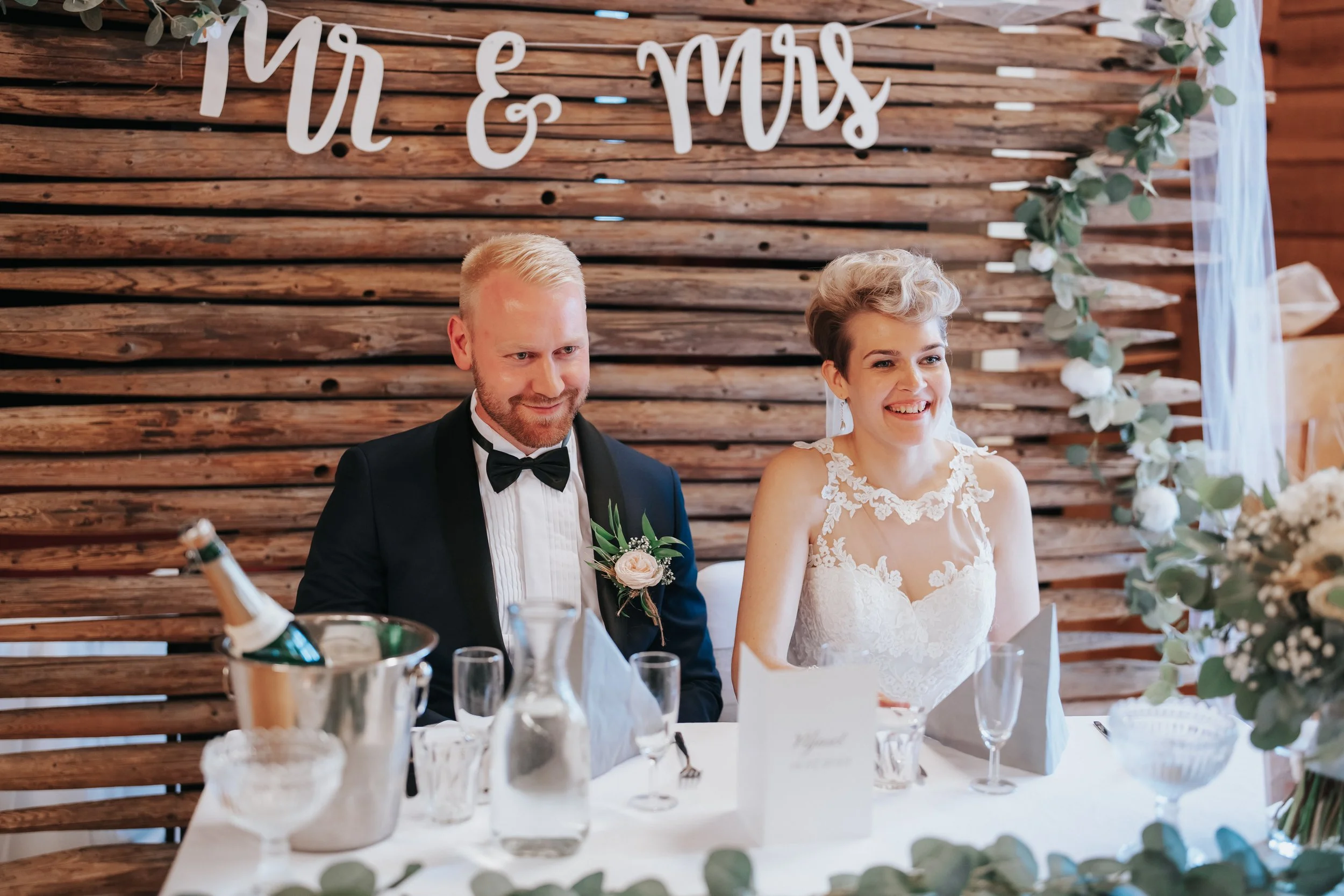 Bride and groom sitting at a wedding table with 'Mr & Mrs' sign and floral decorations.