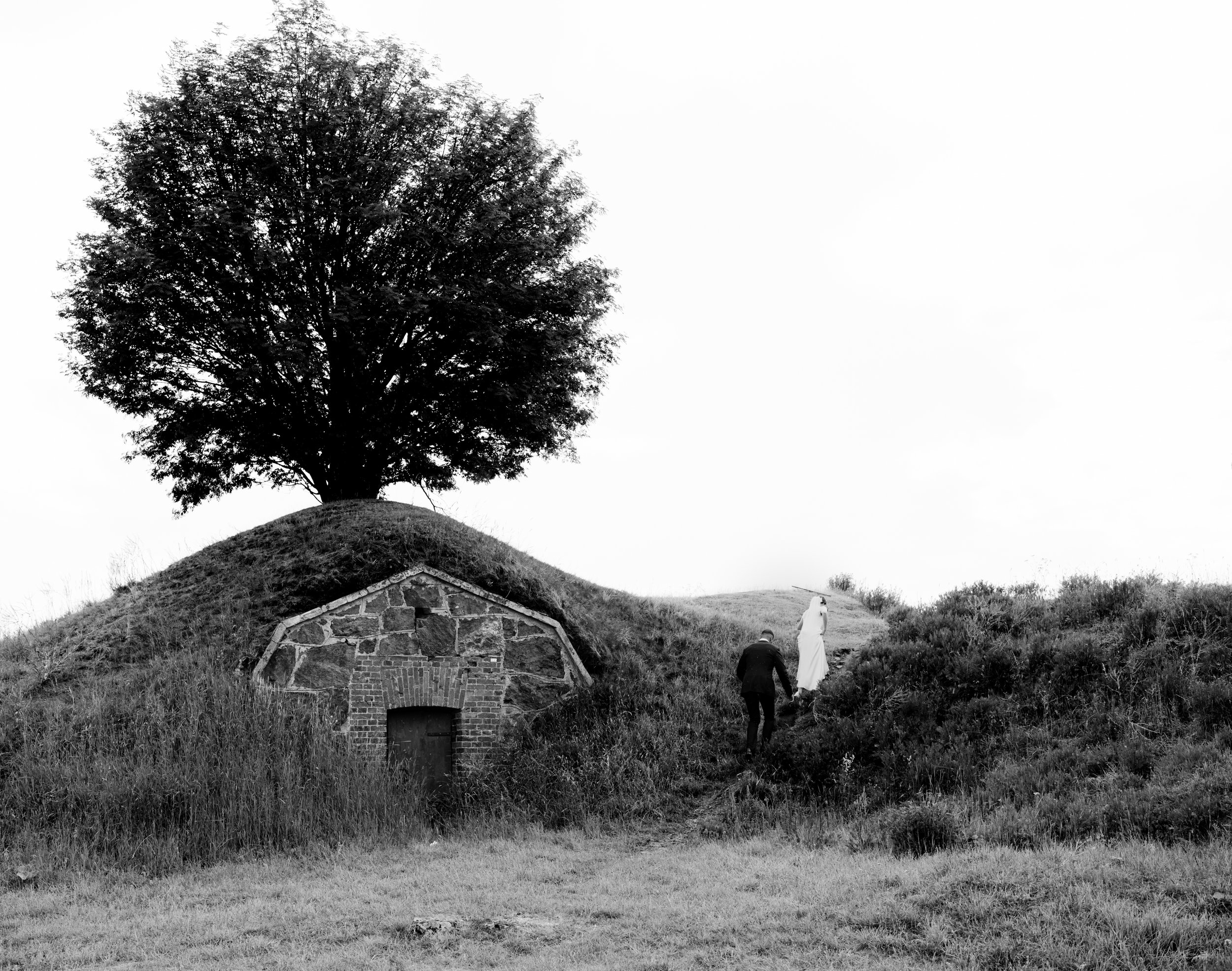 Silhouette of a tree on a grassy mound with a stone structure and door at its base, and a couple walking up the hill.