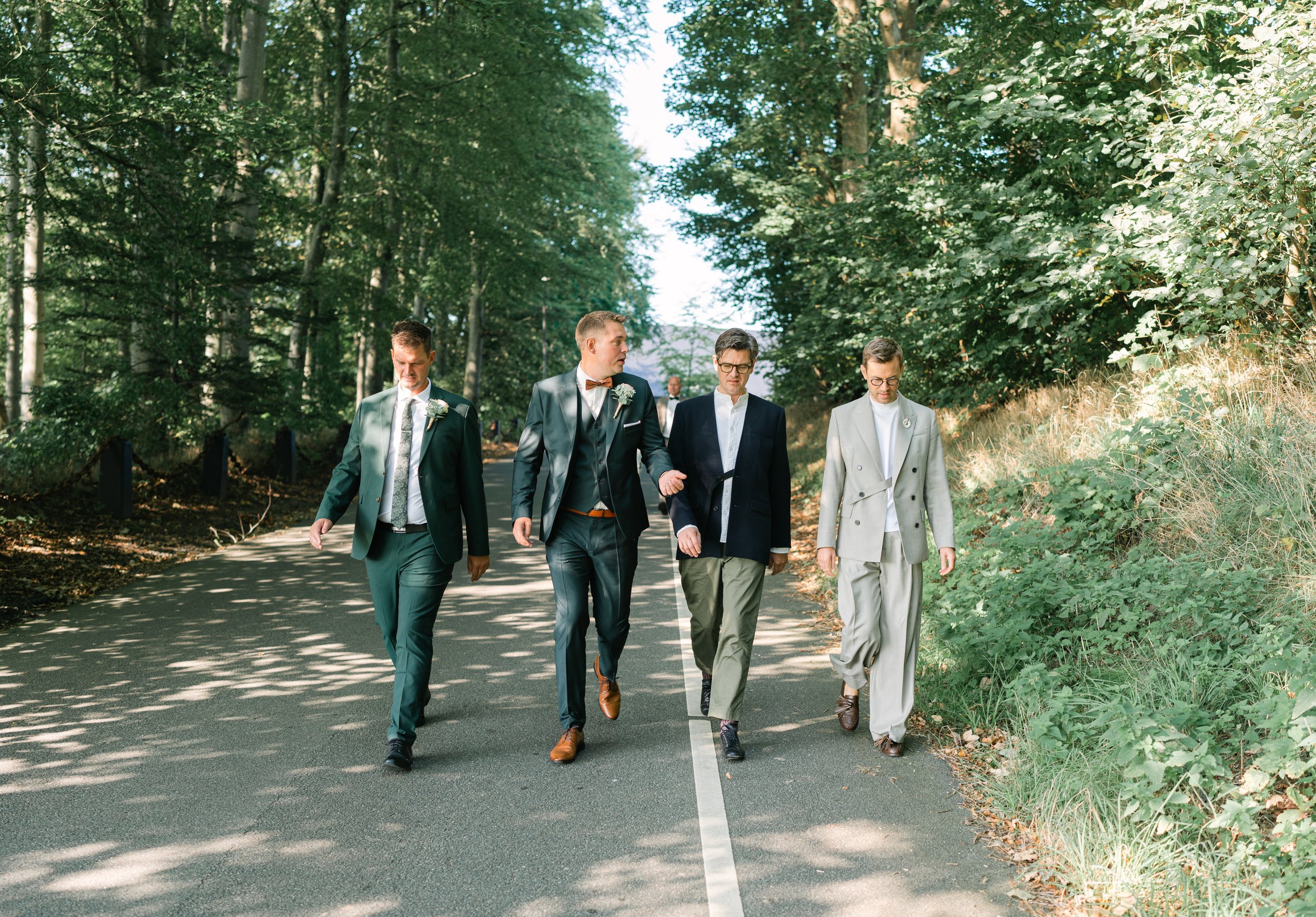 Four men in formal attire walking down a tree-lined road in a forested area.