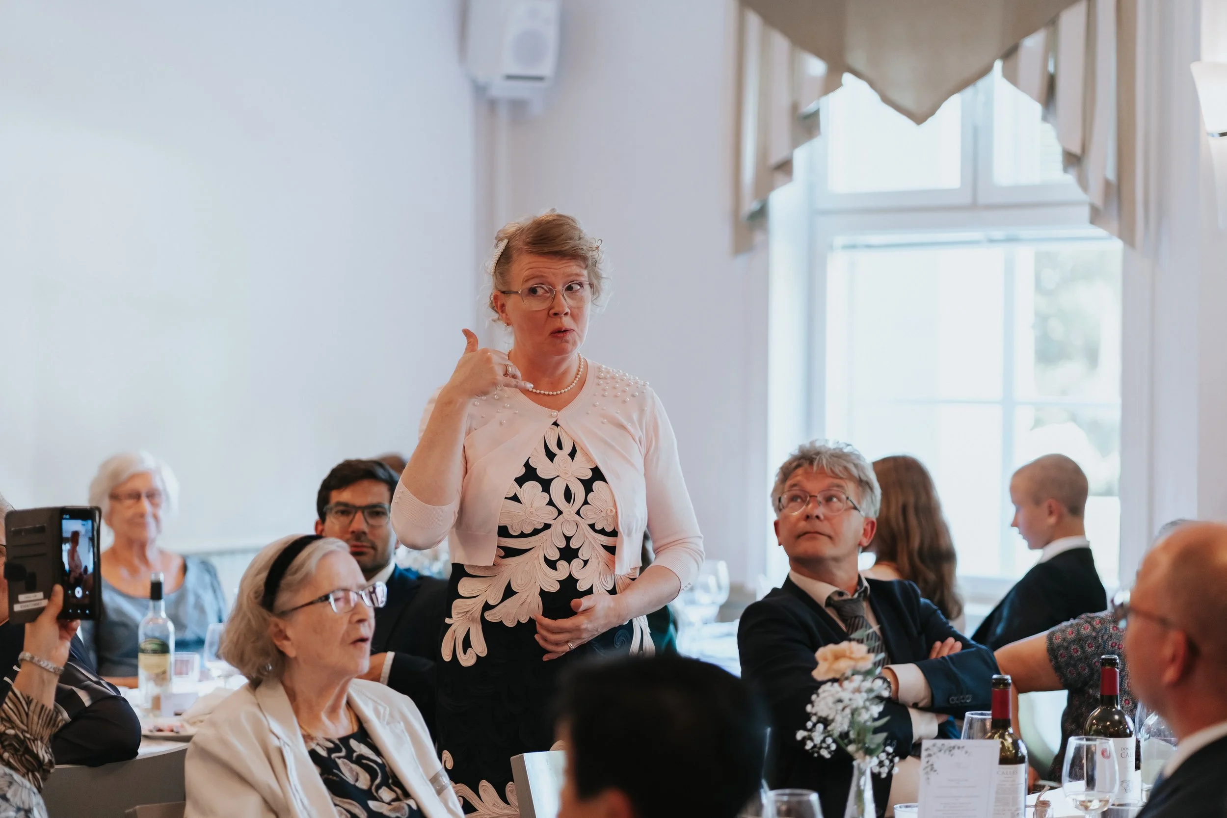 Woman gesturing while speaking at an indoor event, surrounded by seated people.