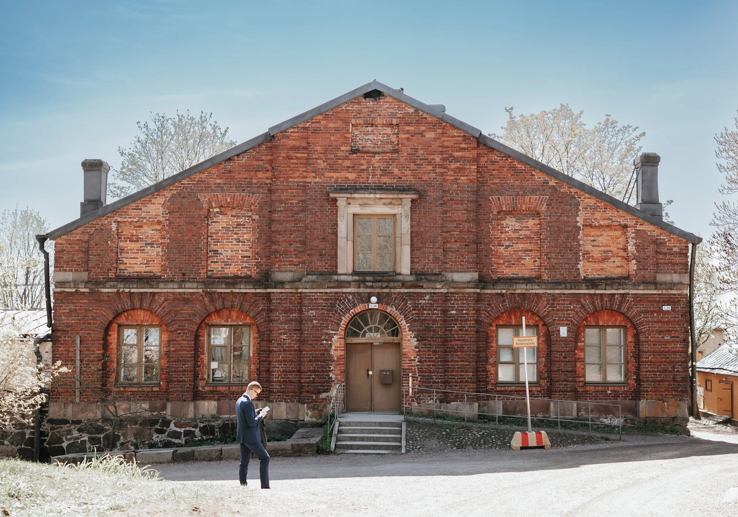 Historic brick building with arched windows and a central entrance, person walking in front holding a phone.