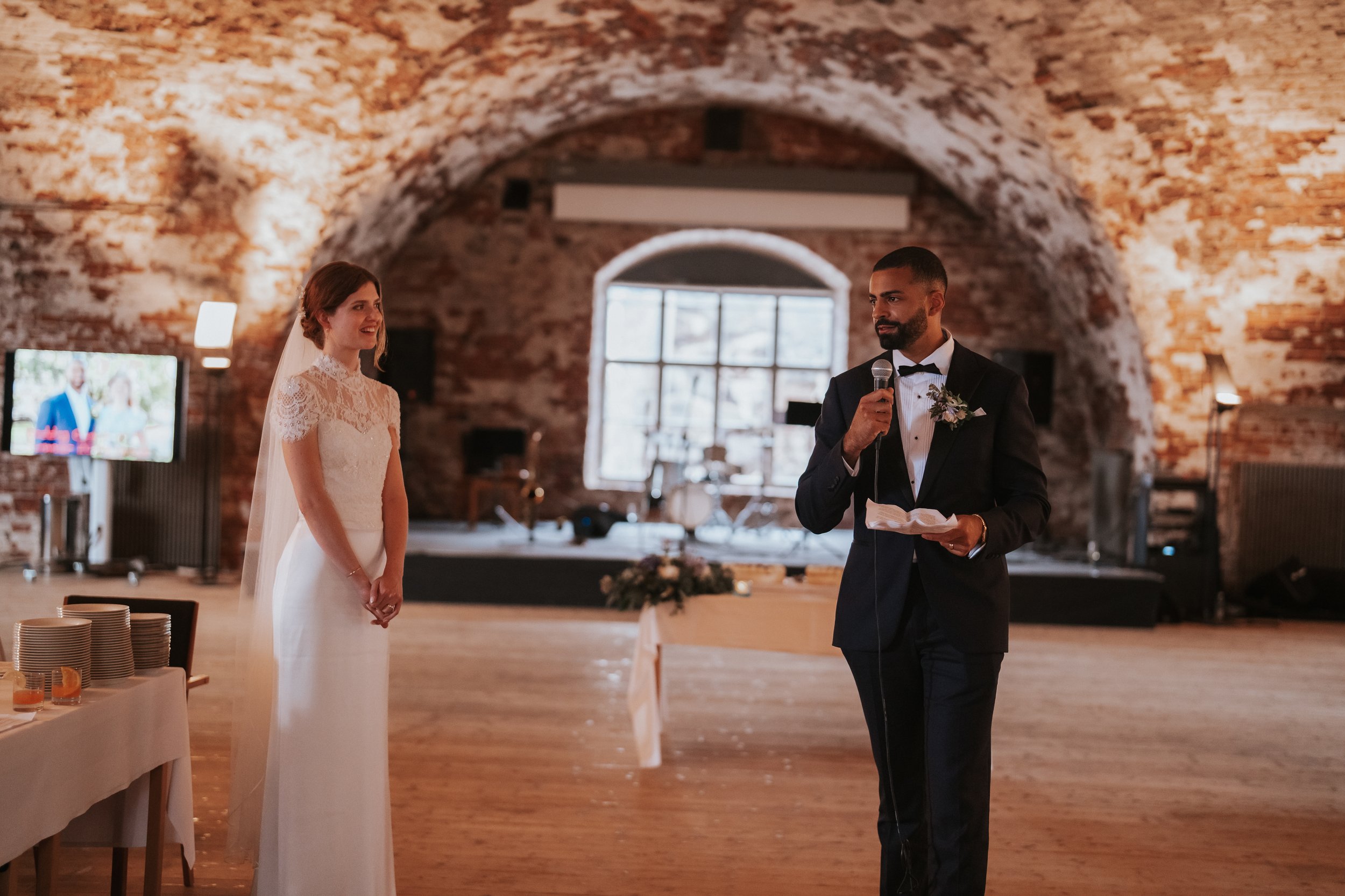Wedding couple in brick venue, bride in white dress, groom in suit giving speech with microphone.
