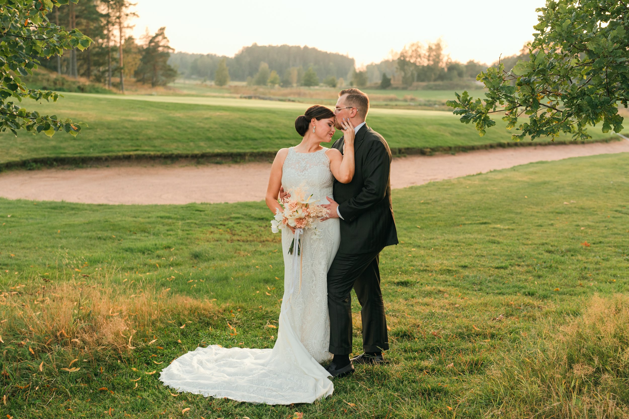 Bride and groom embracing on a grassy field, surrounded by trees, with the bride holding a bouquet and wearing a wedding dress, and the groom in a suit.