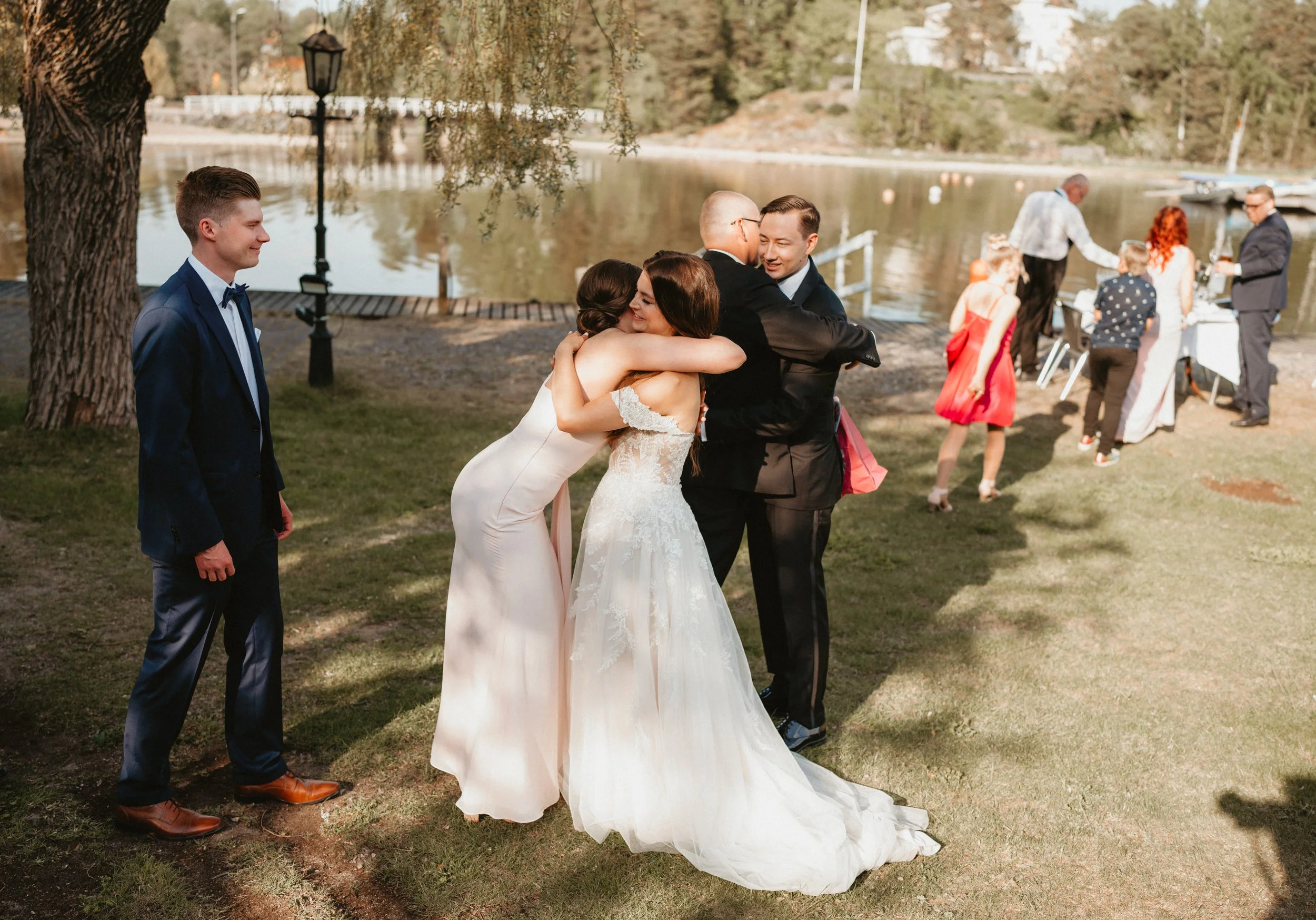 Wedding guests embracing near a lake with trees and outdoor decorations.