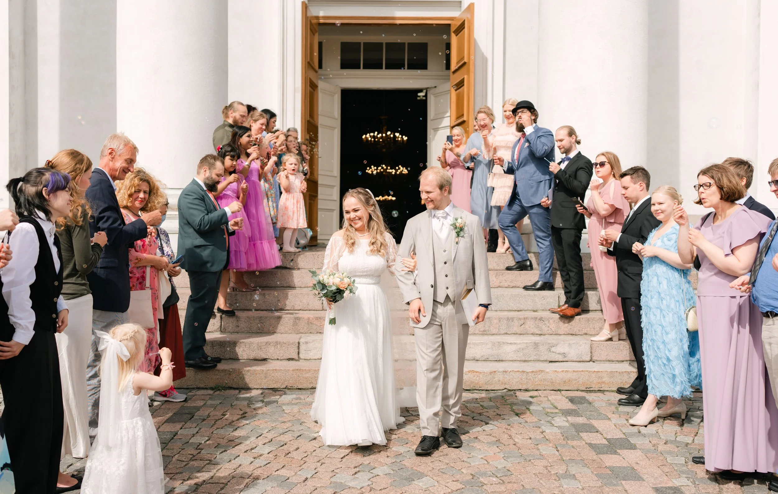 Bride and groom walking down stone steps surrounded by guests celebrating with bubbles. Guests wear formal attire.