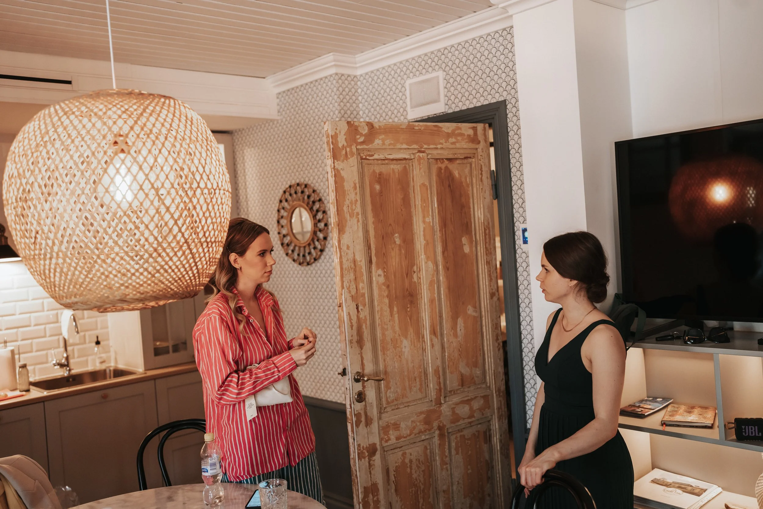 Two women conversing in a stylish interior with a wooden door, patterned wallpaper, decorative mirror, large woven light fixture, and a modern kitchen area.