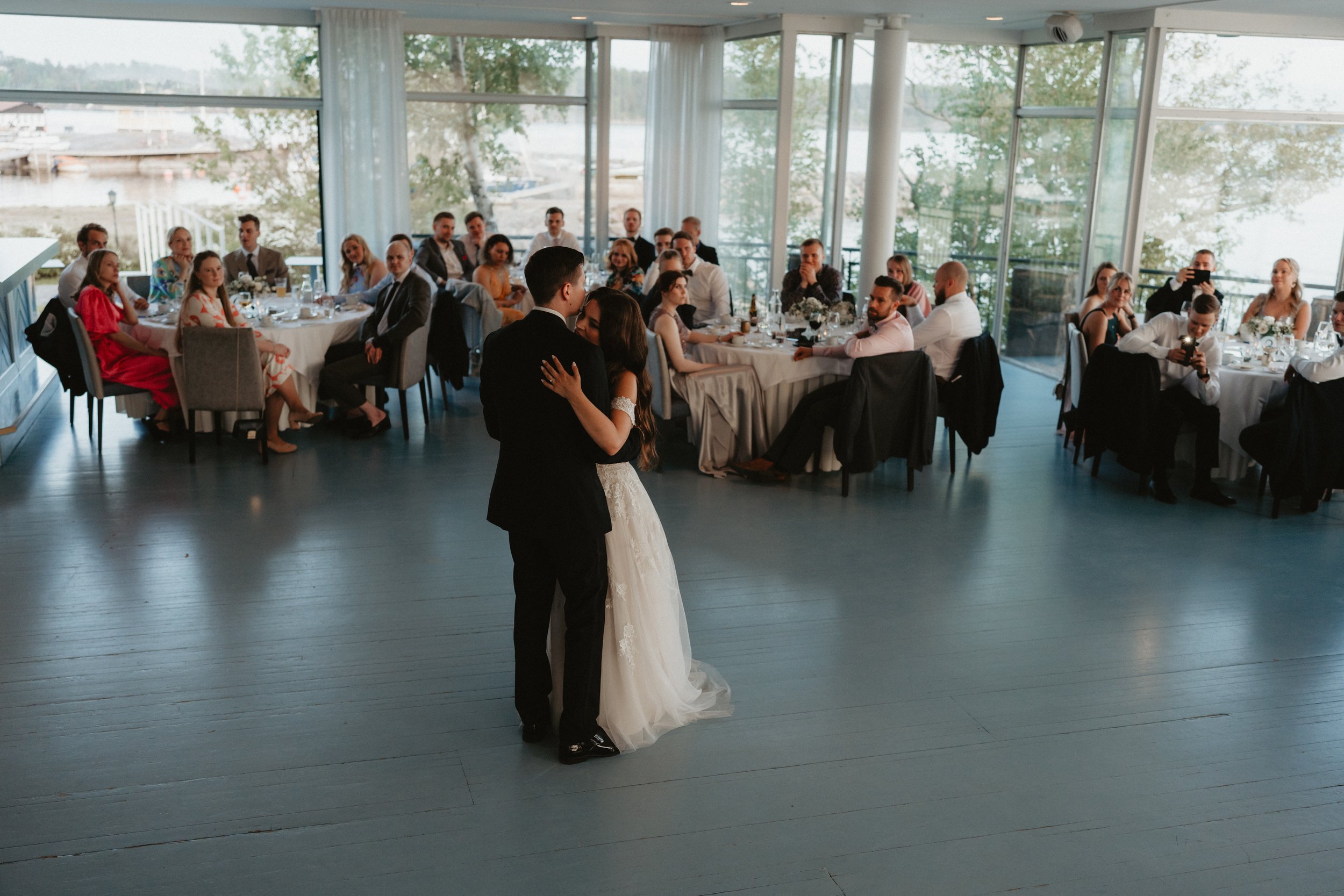 Bride and groom dancing at wedding reception surrounded by guests seated at tables in a venue with large windows.