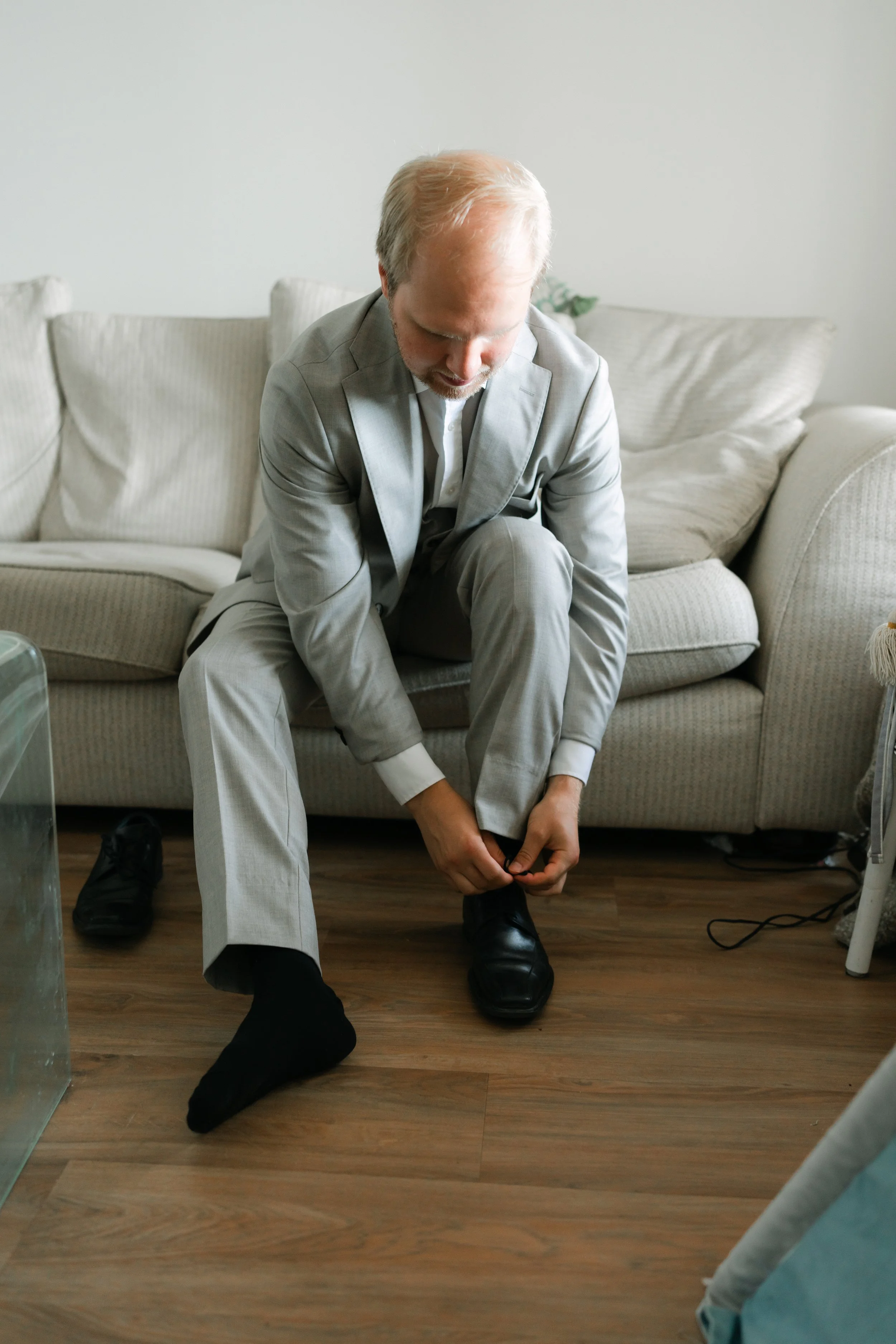 Man in gray suit tying black dress shoe on wooden floor near sofa.
