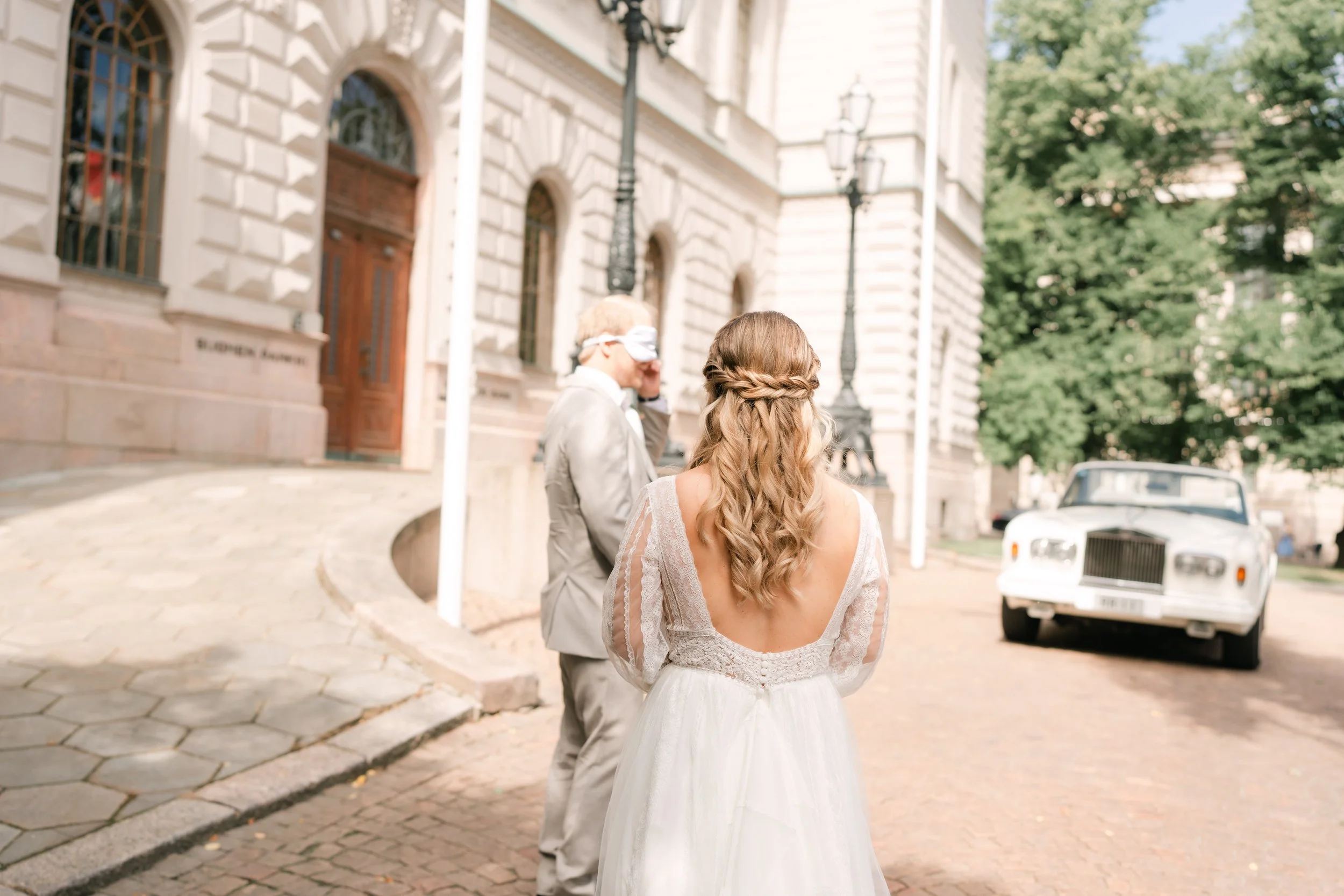 Bride and groom in wedding attire outside historic building with classic car in background.