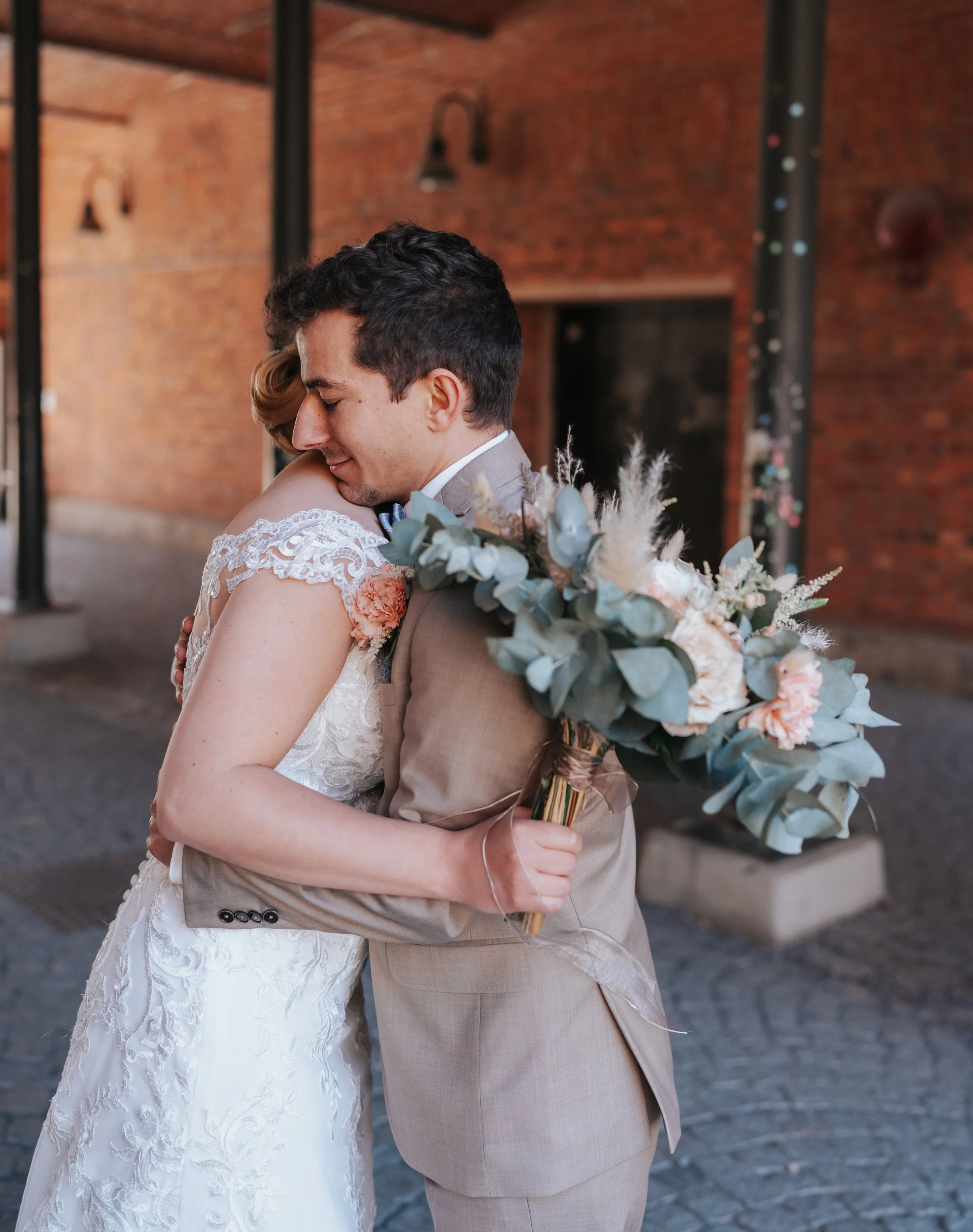 Wedding couple embracing with a bouquet, wearing formal attire in an outdoor setting with a brick wall.
