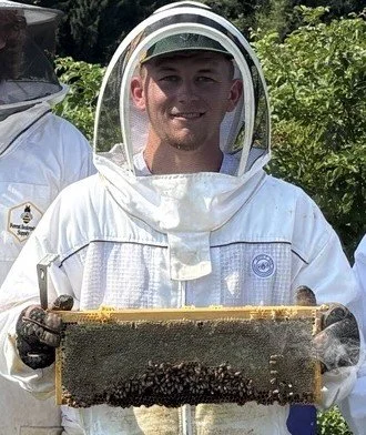 A young woman outdoors with blond hair smiling, wearing a dark jacket and a patterned scarf around her neck, with a blurred background of buildings and sky.