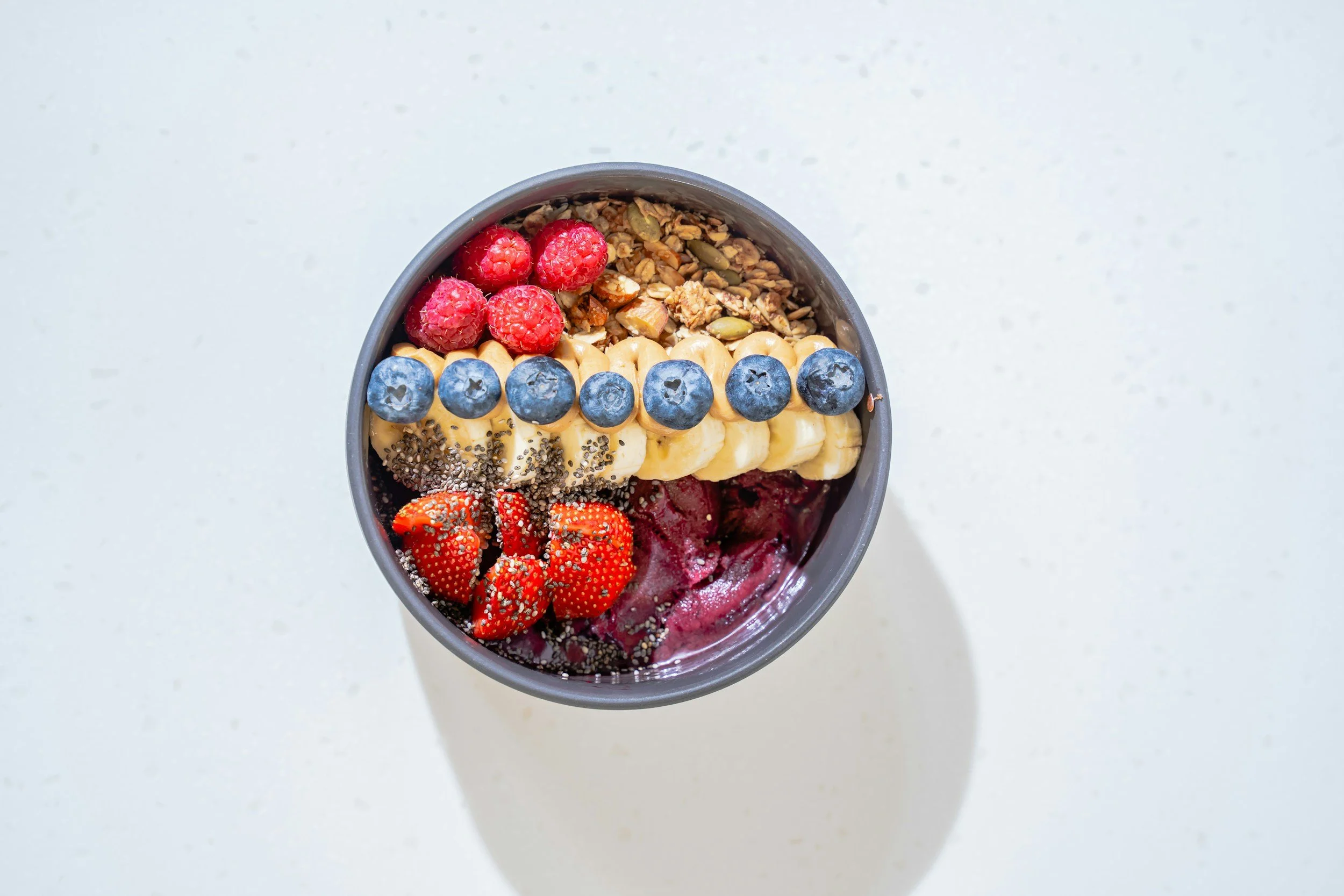 A top view of a bowl containing sliced bananas topped with blueberries, raspberries, strawberries, granola, and chia seeds on a white surface.