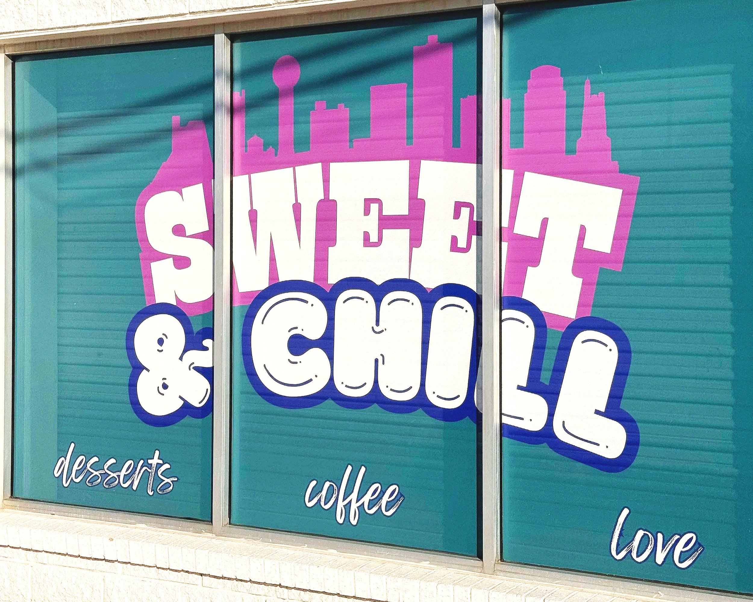 Storefront window with colorful sign promoting sweets, coffee, and love, featuring a pink city skyline silhouette in the background.