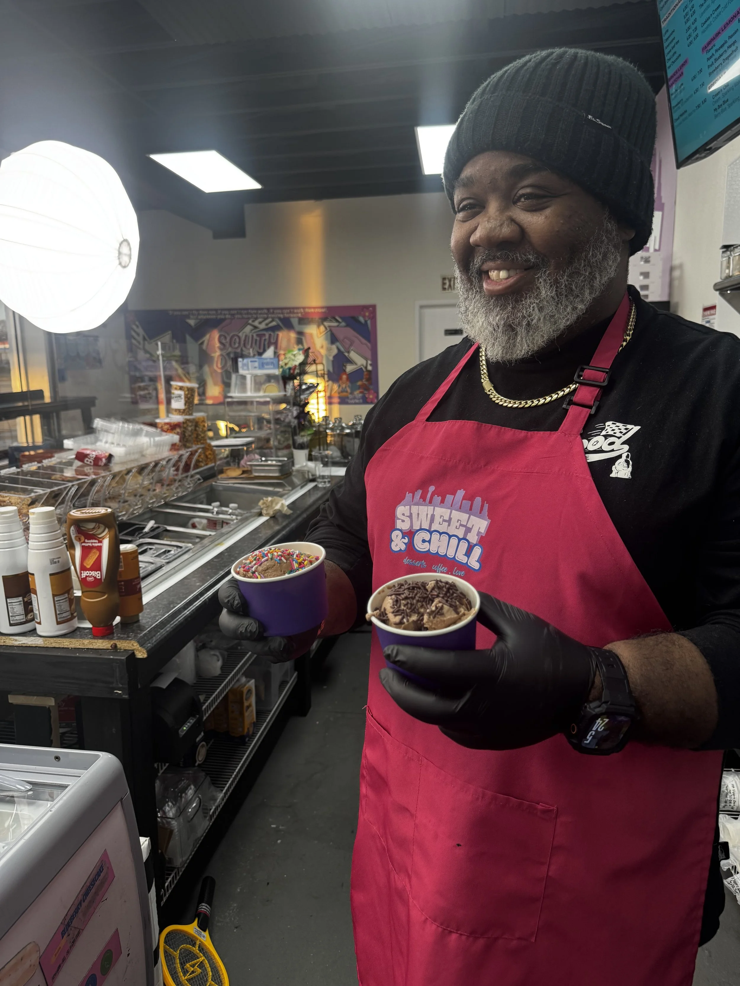 A man with a beard and sunglasses wearing a white shirt and red apron with a logo that reads 'Sweet & Chill' in a colorful ice cream shop.