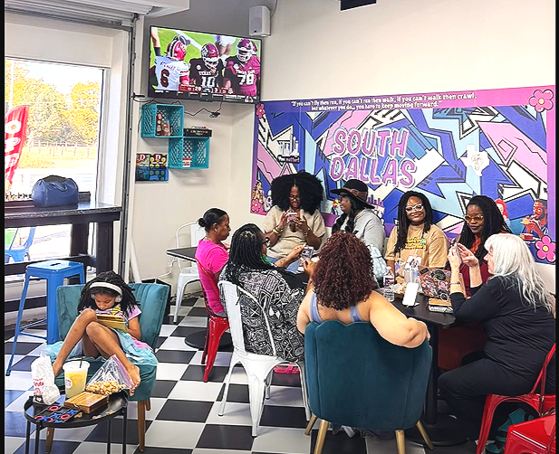 A group of women and a girl sitting at a table in a colorful restaurant decorated with graffiti art and South Dallas signs, watching a football game on TV.