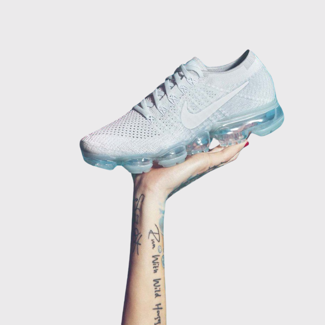 A hand with tattoos holding a white Nike air vaporMax running shoe against a plain gray background.