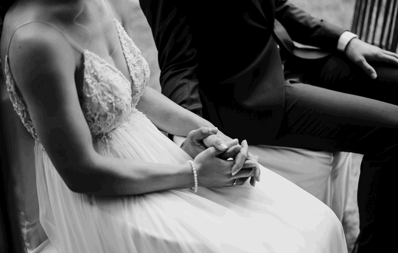 Ein Paar sitzt in formeller Kleidung; die Frau trägt ein Hochzeitskleid und der Mann einen Anzug. Sie halten sich an den Händen. Schwarz-Weiß-Fotografie.