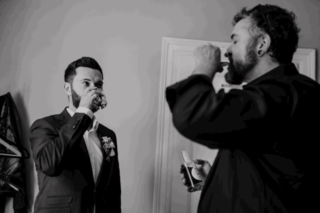 Zwei Männer trinken in einem Raum, der eine trägt einen Anzug und Blumenschmuck, der andere Freizeitkleidung. Schwarz-weiß Fotografie.