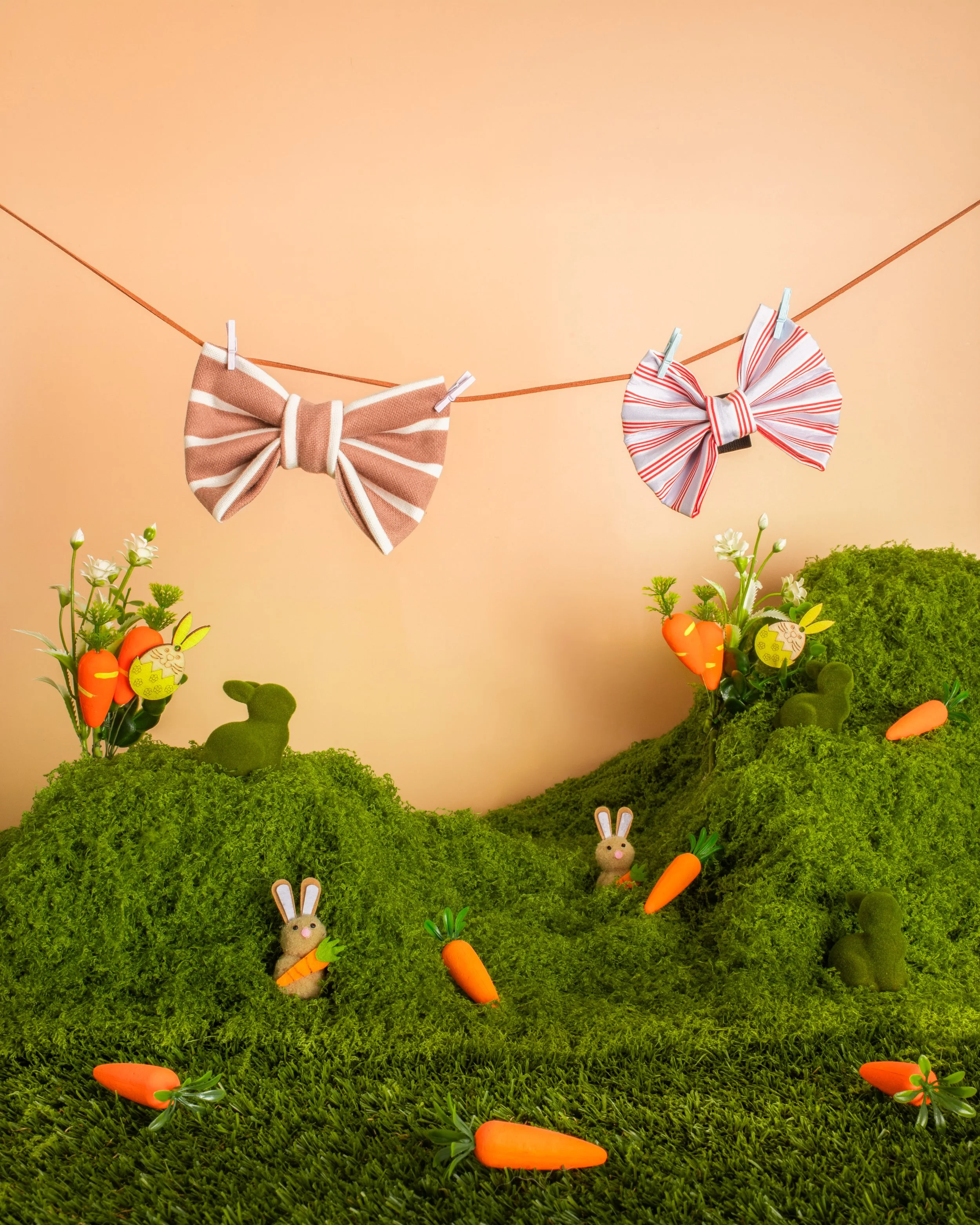 Decorative scene with felt bunnies holding carrots, hanging on a string with clothespins, set against green moss and small plants, on a peach background.