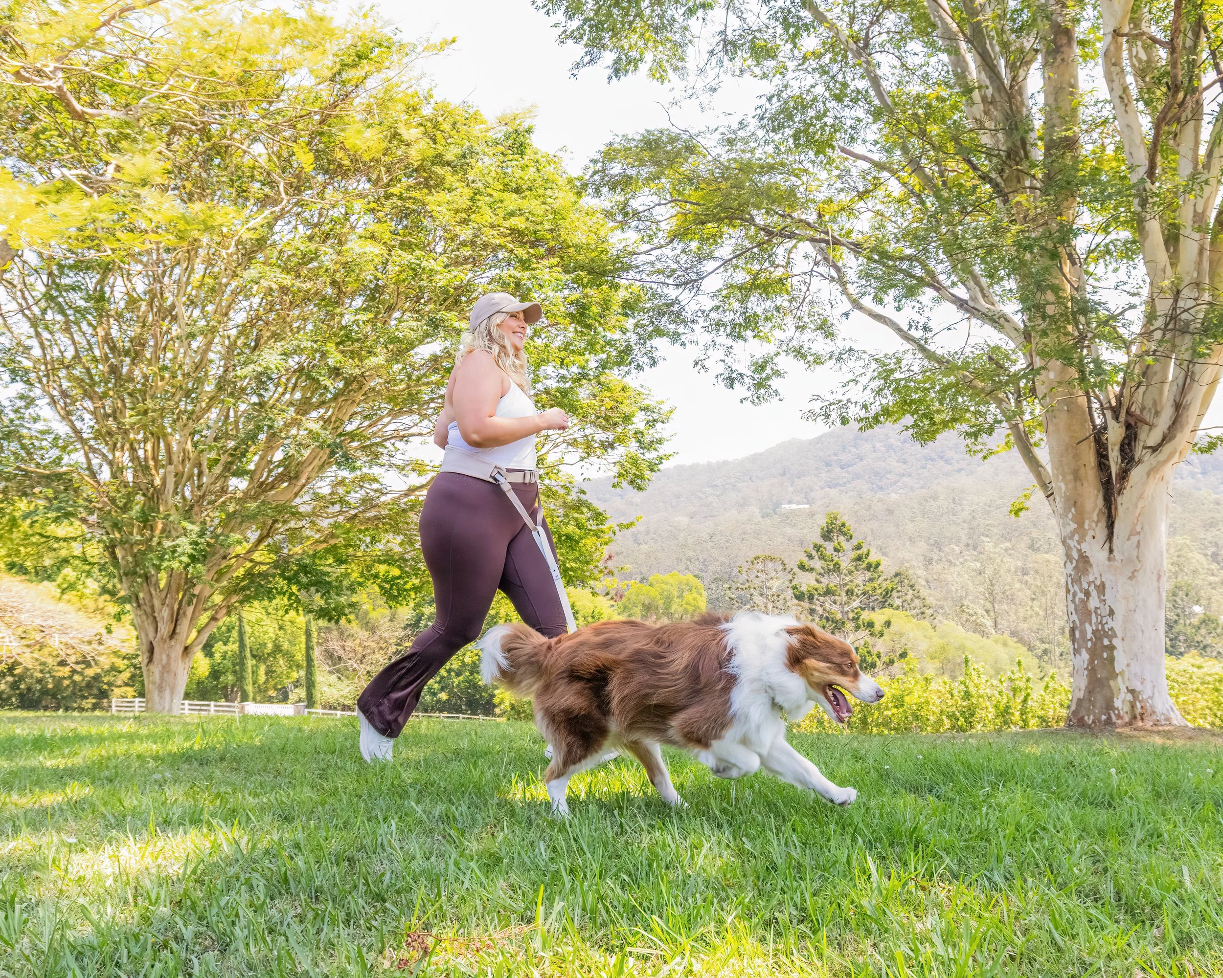 A woman in athletic clothing walking her brown and white dog on a leash in a park with trees and green grass.