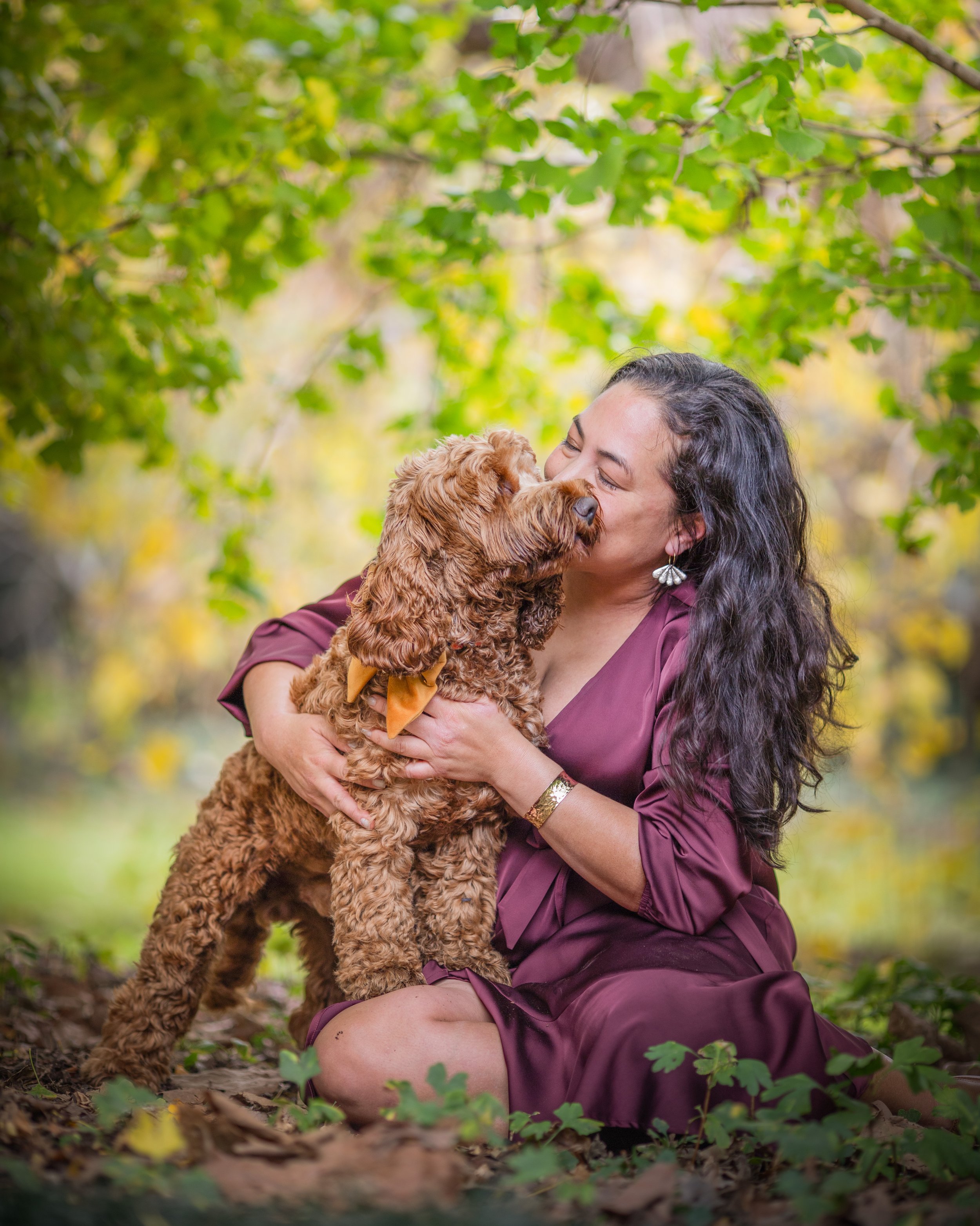 Woman in purple dress hugging and playing with a curly-haired brown puppy in a park with green leaves and autumn colors.