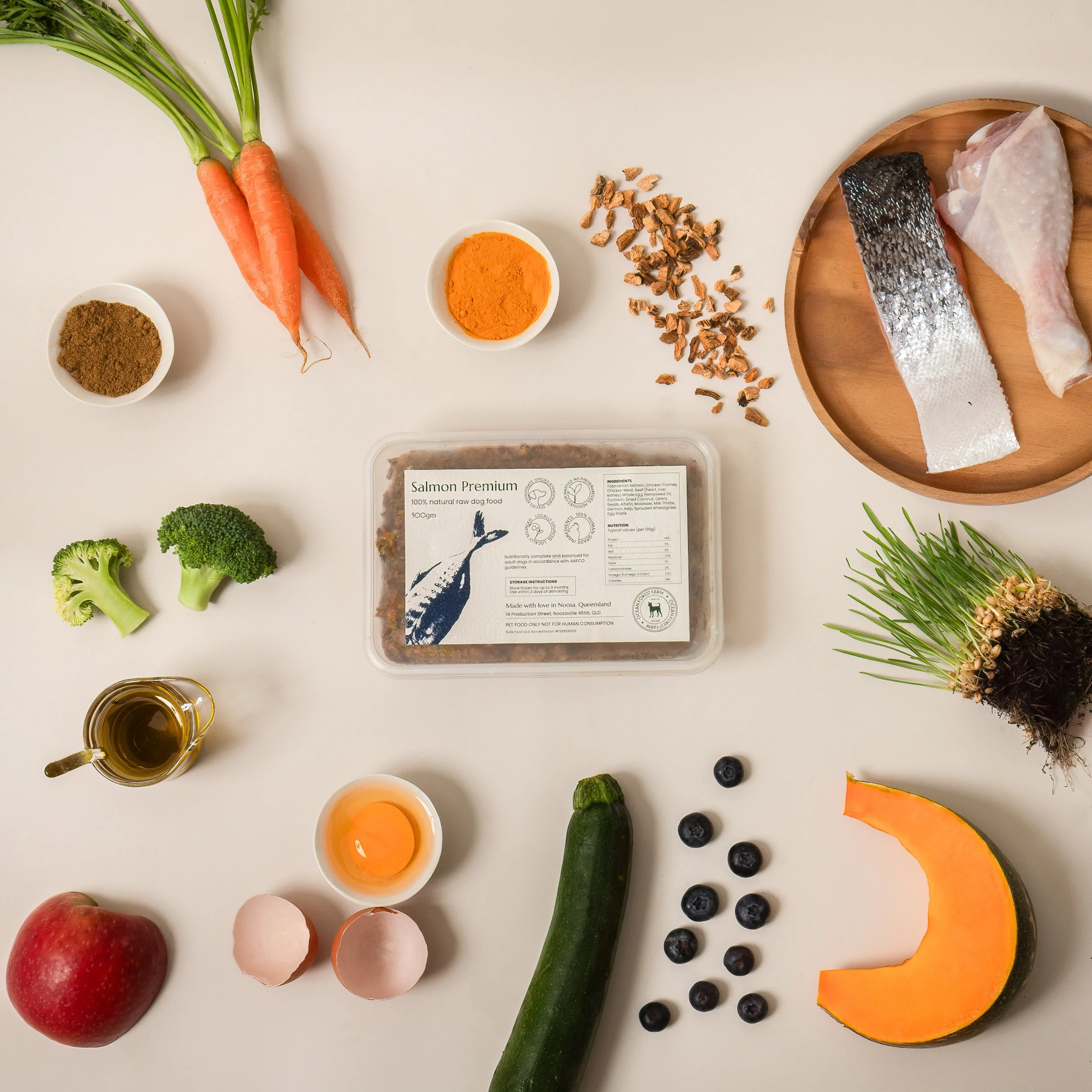 Assorted fresh vegetables, fish, eggs, spices, and blueberries on a white surface, with label indicating organic salmon pet food.